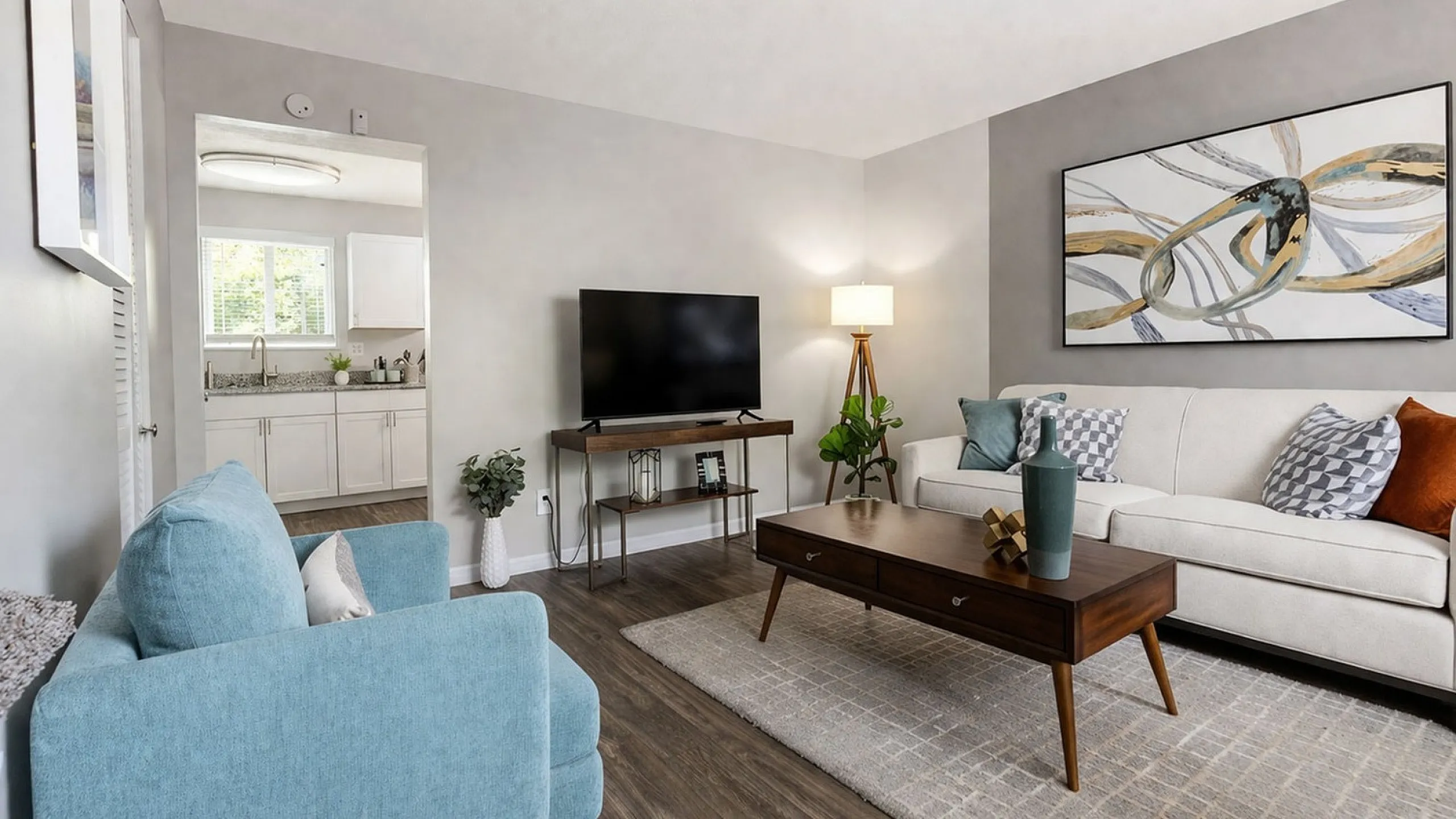Modern living room with stylish decor, featuring a plush white sofa, sleek TV stand, and vibrant abstract artwork. Open view into a bright, well-equipped kitchen.