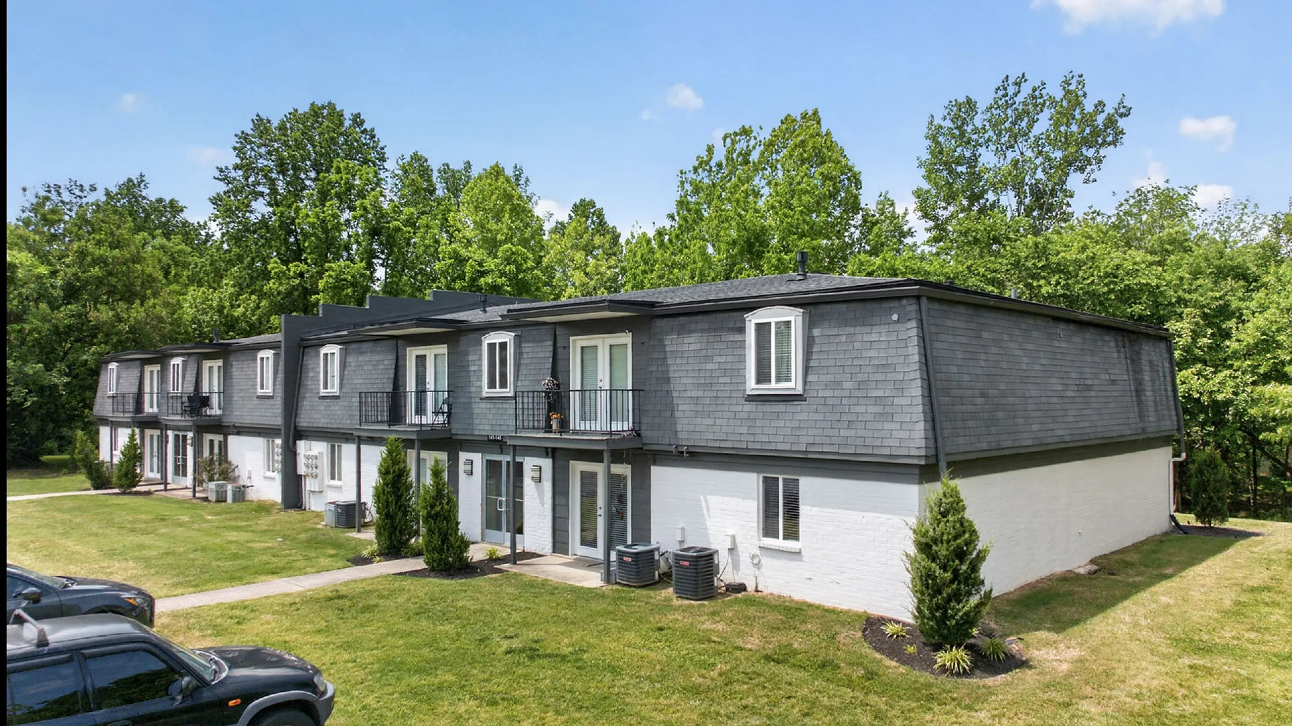 Charming townhouse complex with modern gray exteriors, lush green lawns, and inviting balconies against a backdrop of vibrant trees.