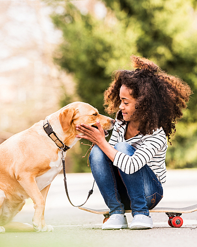 Joyful young girl bonding with her Labrador while sitting on a skateboard in a sunny, lush green park.