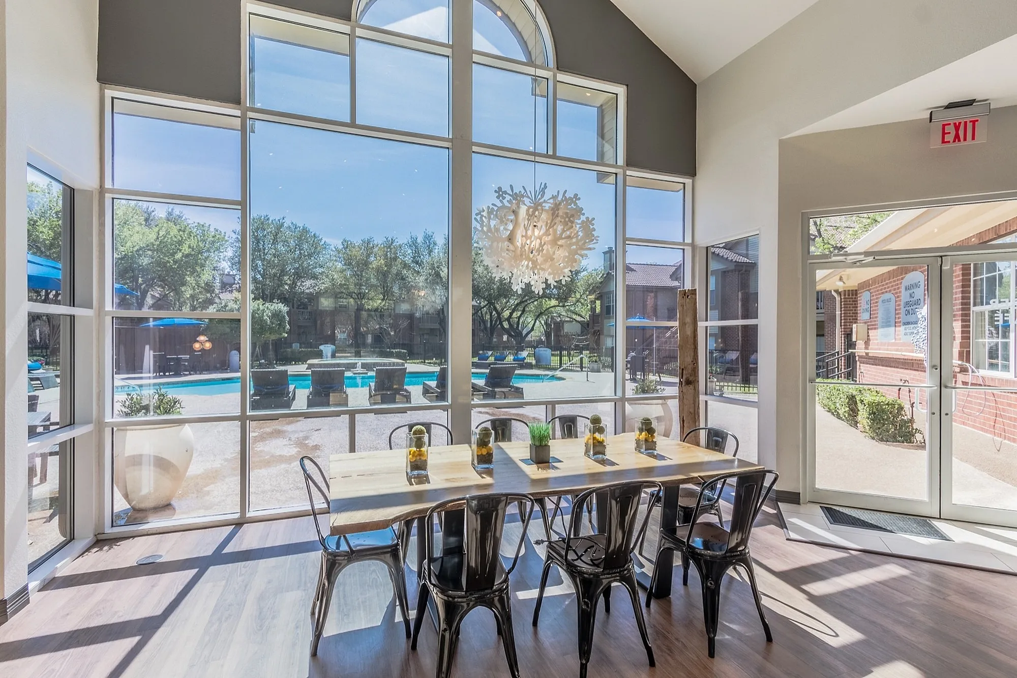 Bright and inviting dining area with large windows overlooking a luxurious pool, featuring modern metal chairs and a rustic wooden table.