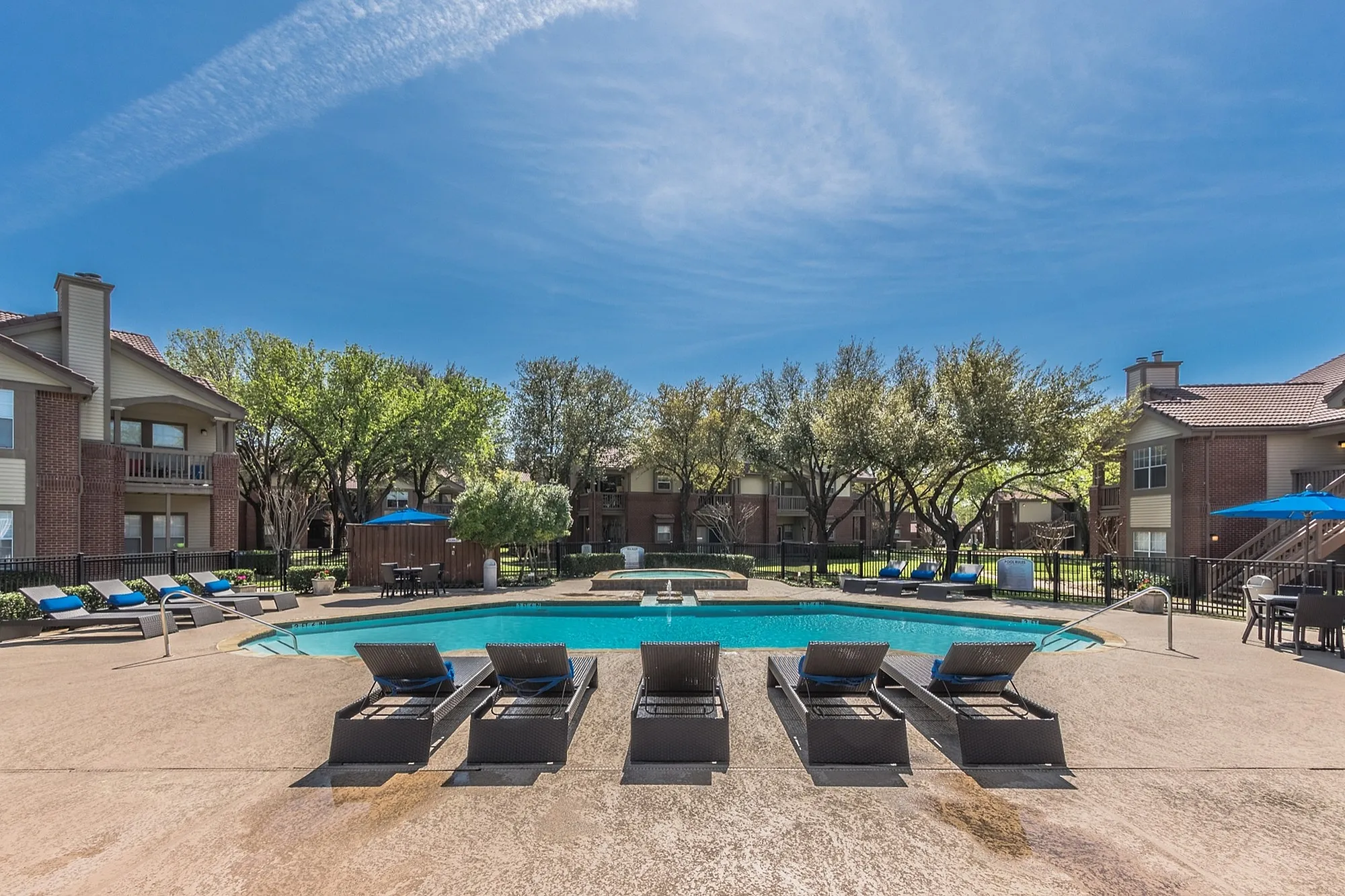 Luxurious apartment complex featuring a sparkling pool with stylish loungers and a serene landscape under a clear blue sky.