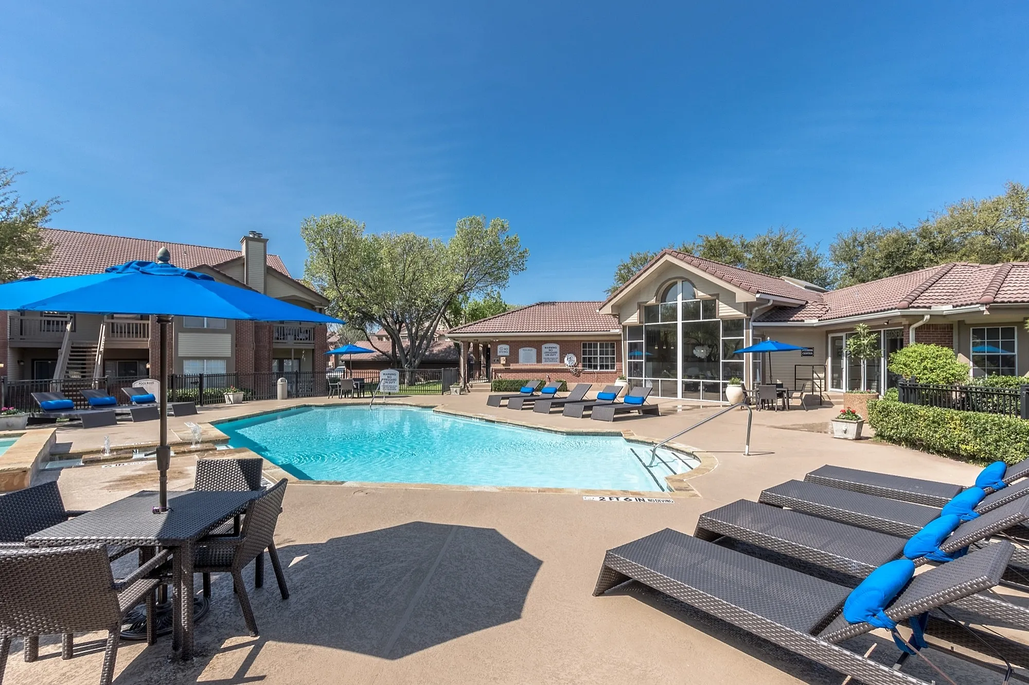 Charming apartment complex featuring a sparkling pool with luxurious lounge chairs, shaded seating areas, and vibrant blue umbrellas, perfect for relaxation and leisure.