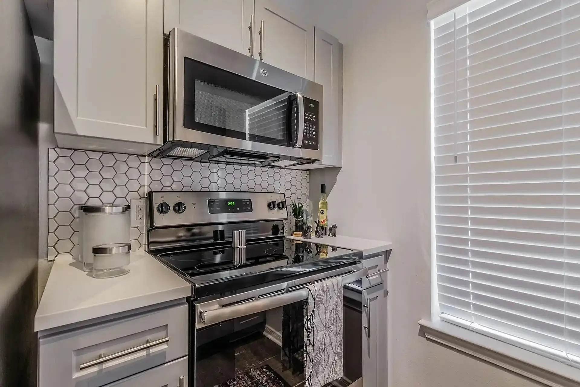 Modern kitchen featuring sleek stainless steel appliances and stylish hexagon tile backsplash, enhanced by ample natural light from large window blinds.