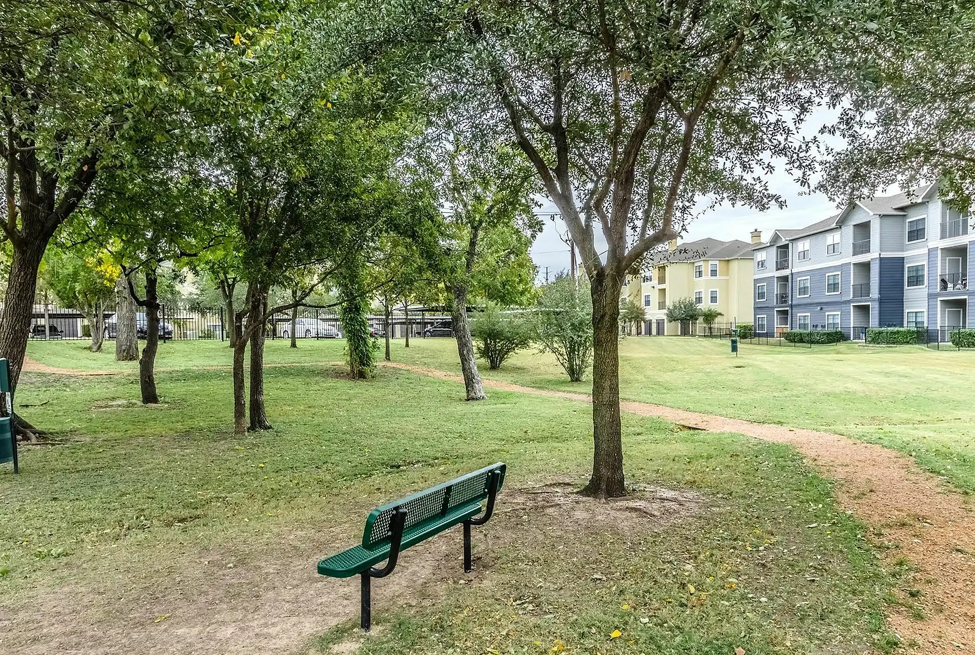 Tranquil park setting with lush greenery, a walking path, and nearby modern apartments, perfect for relaxation and enjoying nature.