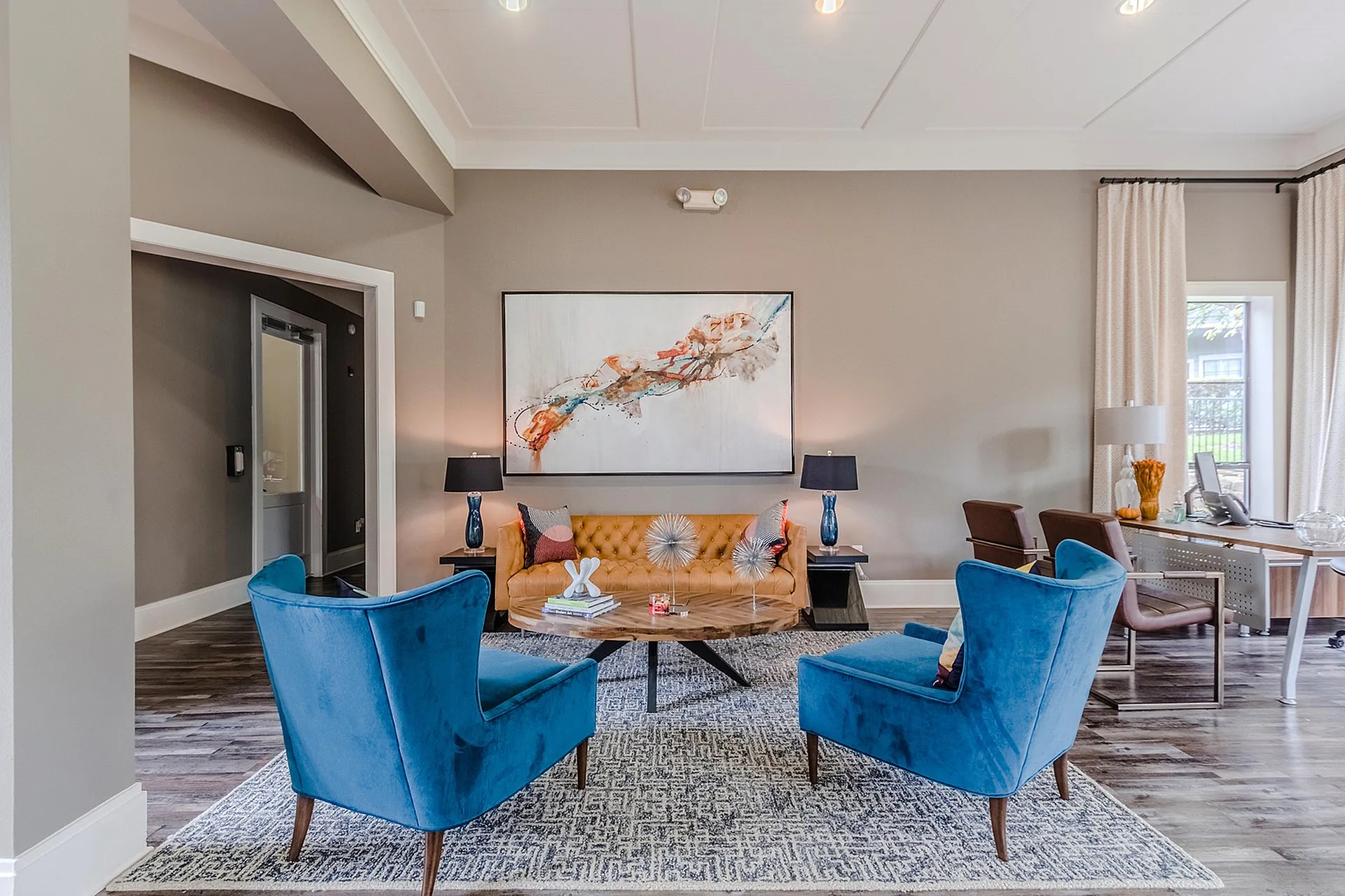 Elegant living room with vibrant blue armchairs, a stylish orange sofa, and contemporary artwork, featuring an inviting workspace by the window.