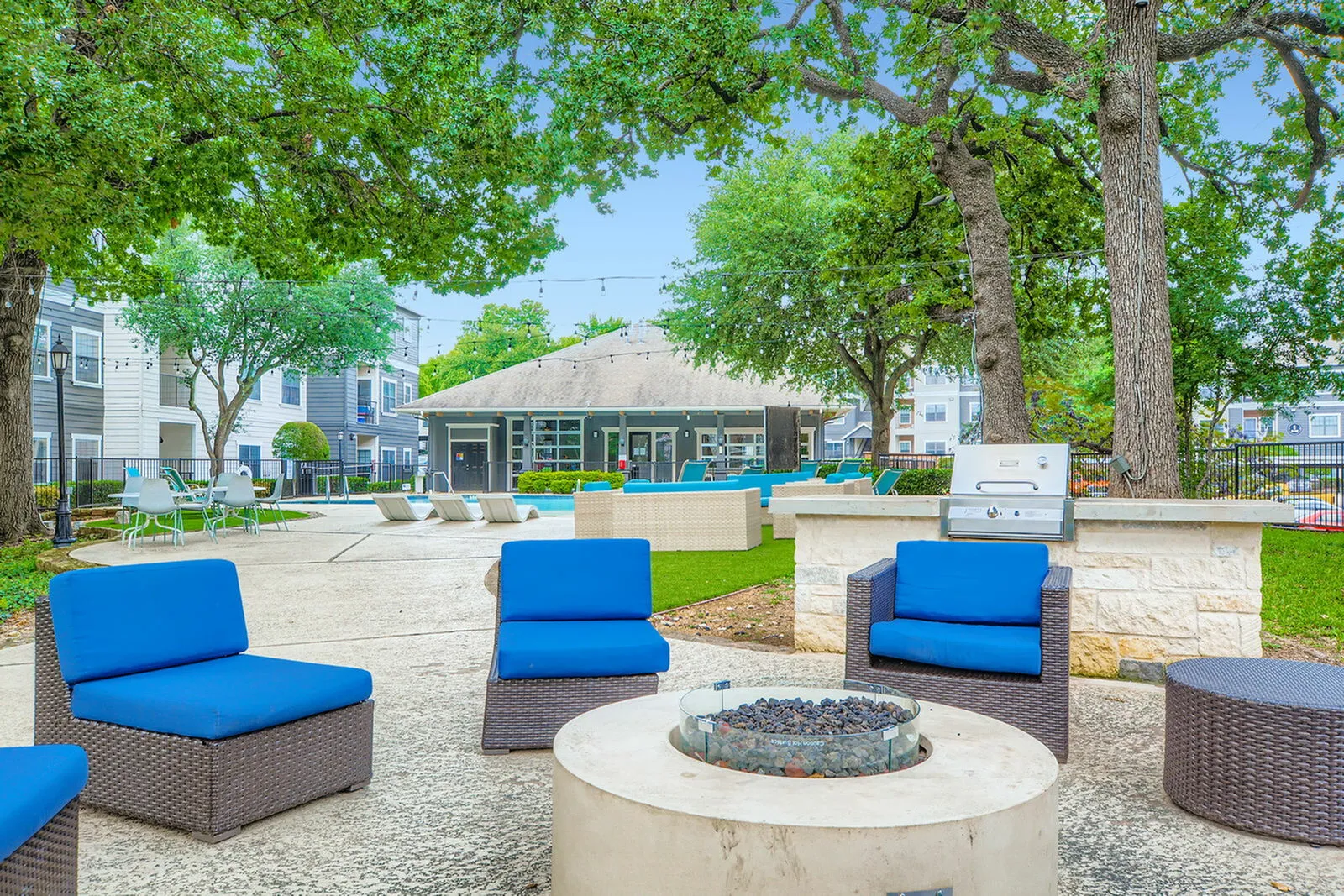 Inviting outdoor lounge area with cozy blue seating, fire pit, and barbecue grill, set amidst lush greenery and a modern clubhouse backdrop.
