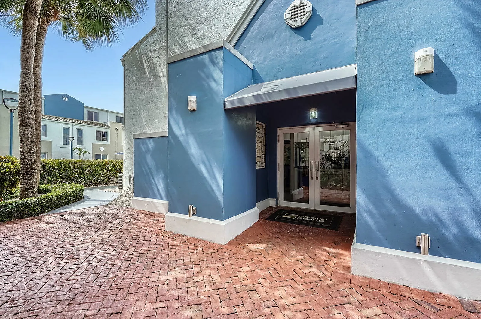 Inviting apartment entrance with vibrant blue walls, surrounded by lush greenery and stylish brick pathway.