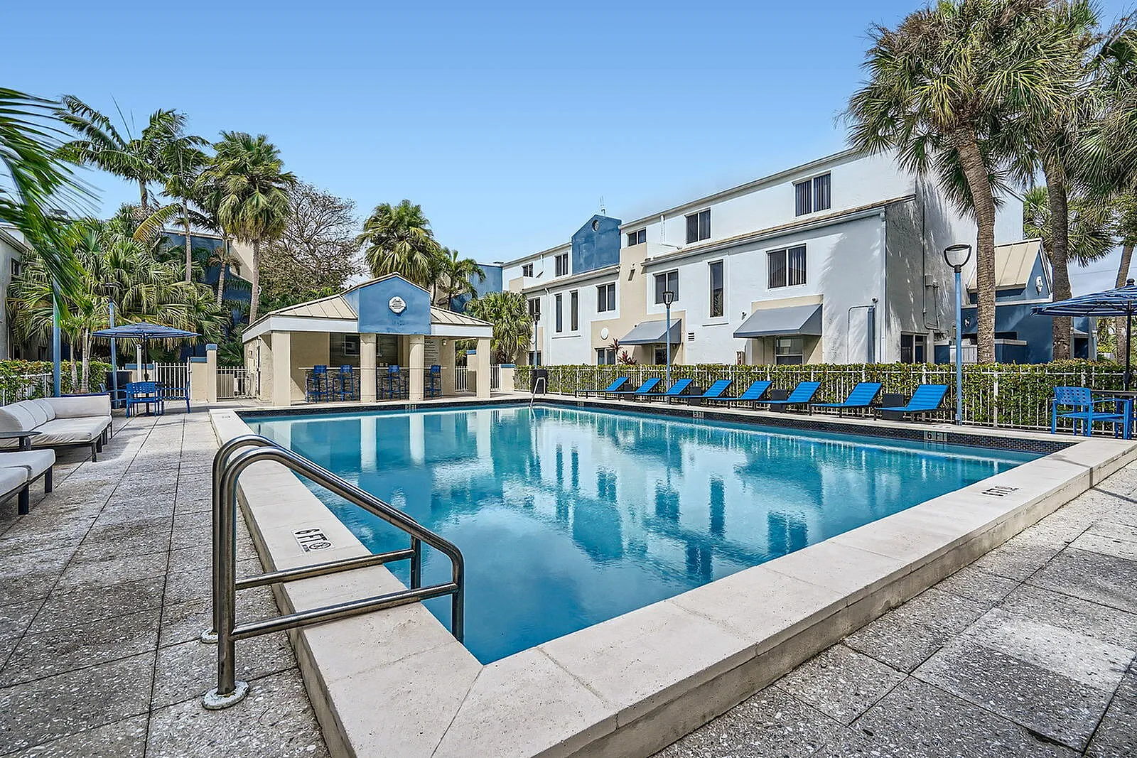 Inviting apartment complex with a crystal-clear swimming pool, lush palm trees, and elegant poolside seating, perfect for relaxation and leisure.
