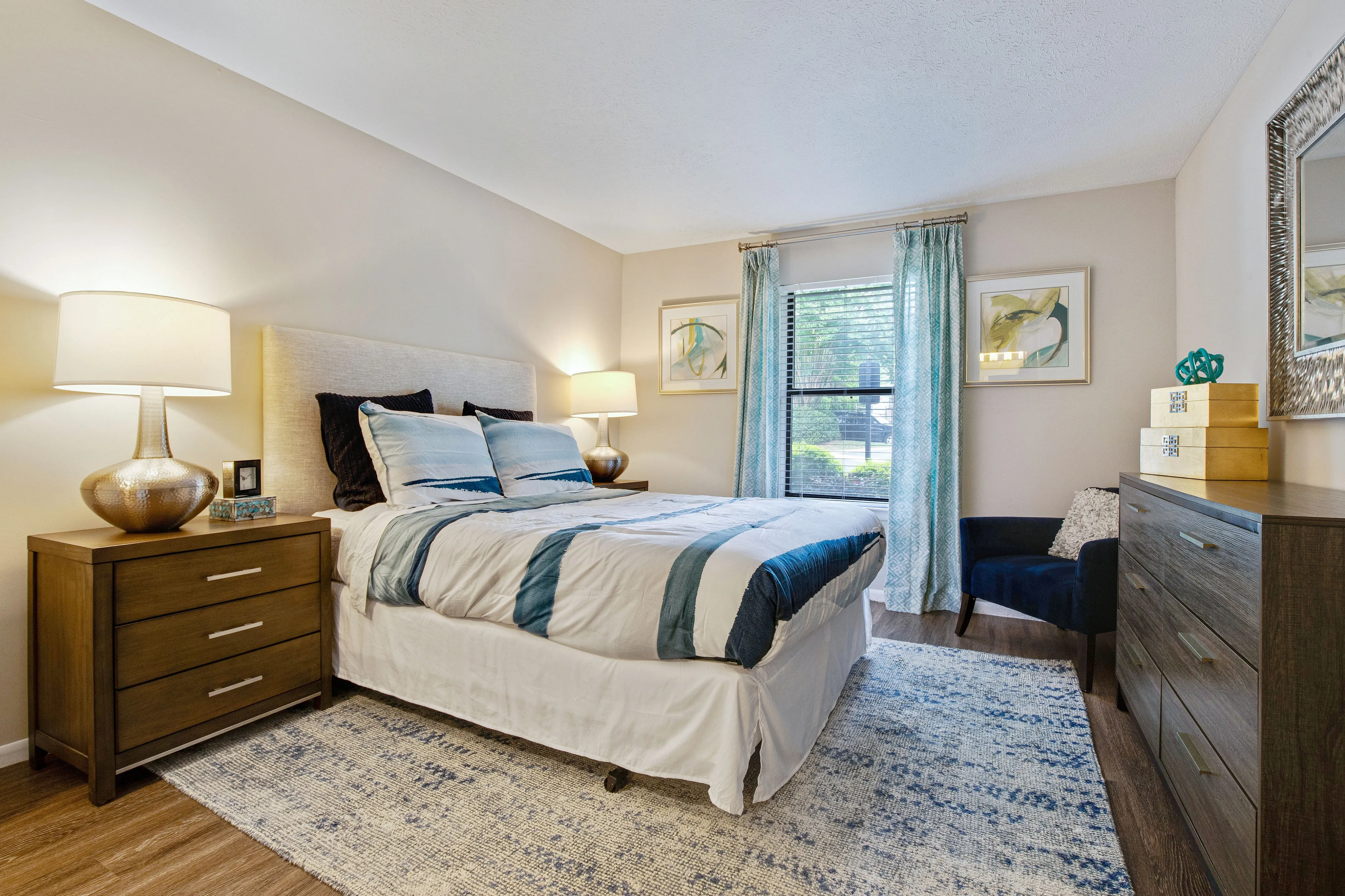 A bedroom with a bed, two side tables with lamps on them, a chest of drawers, a large mirror, a chair, and a window with curtains at The Madison Charlotte, NC apartment rentals.