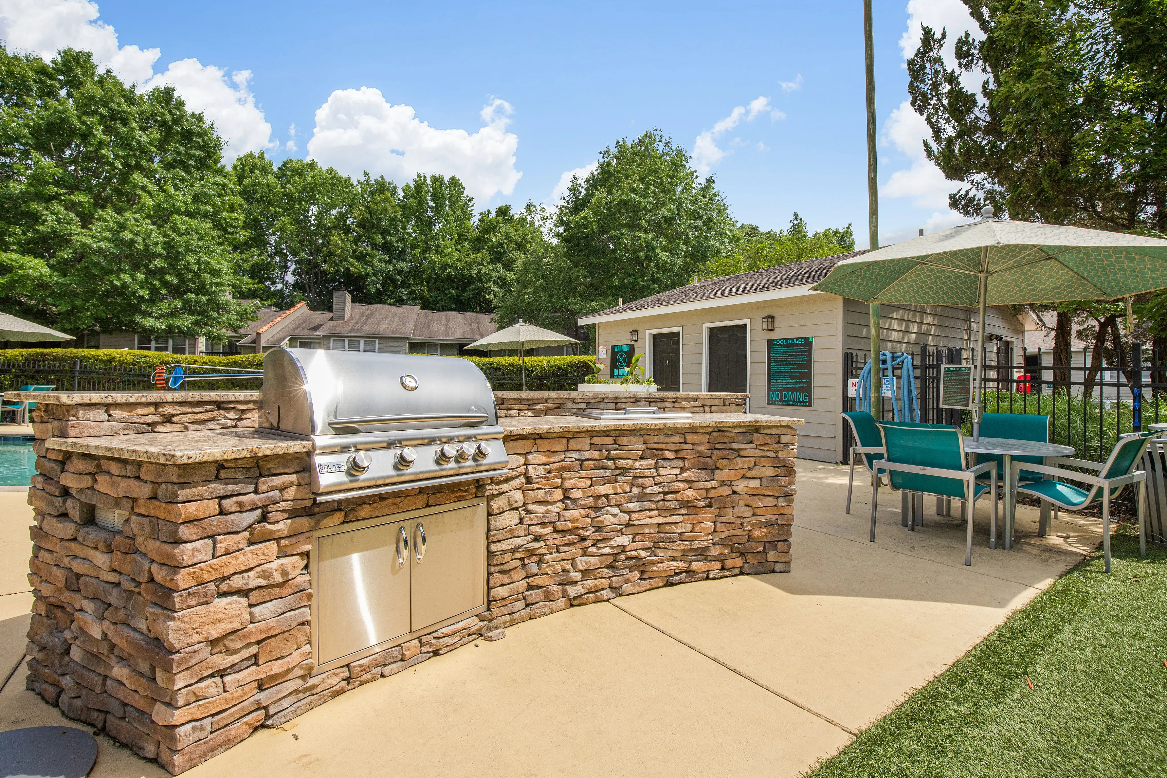 Luxury outdoor grilling area by the pool with stylish stone design and modern barbecue equipment, surrounded by lush greenery and comfortable seating.