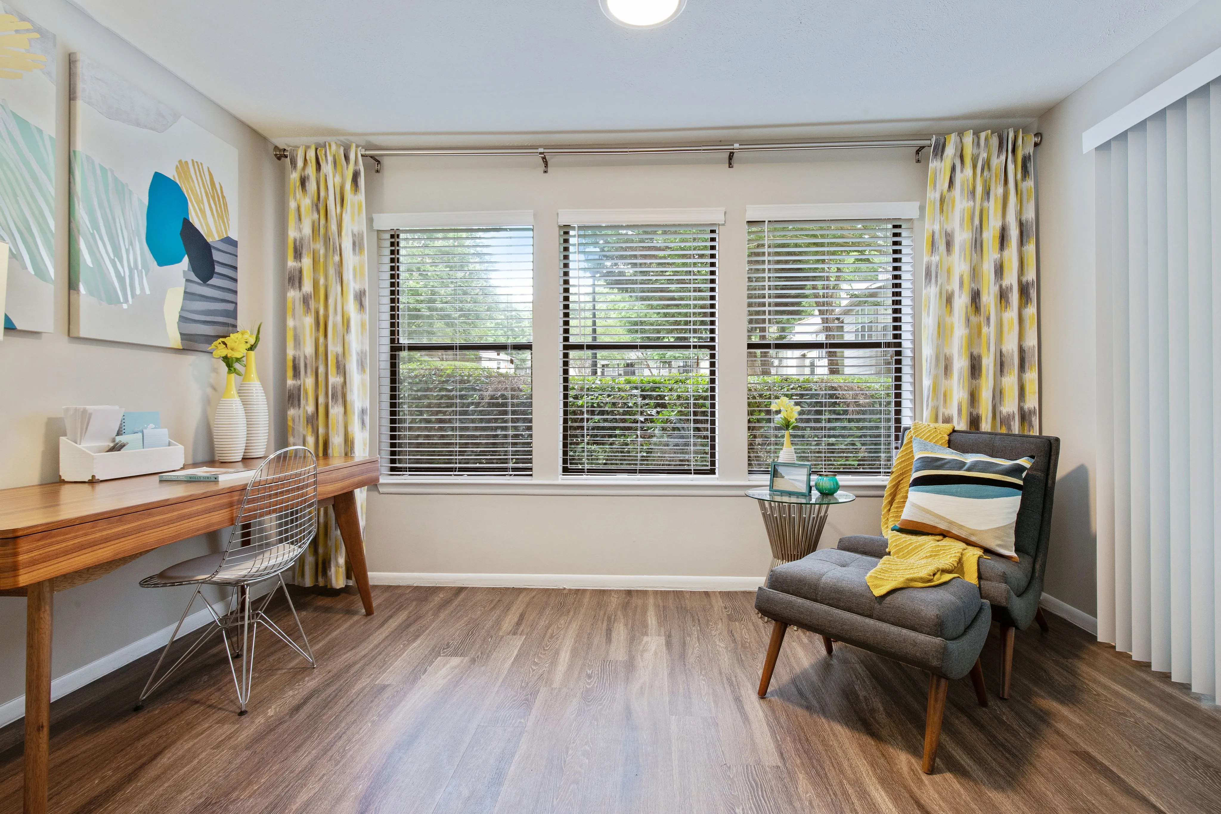 Bright and inviting room with modern decor, featuring a sleek wooden desk and a cozy armchair by large sunlit windows.