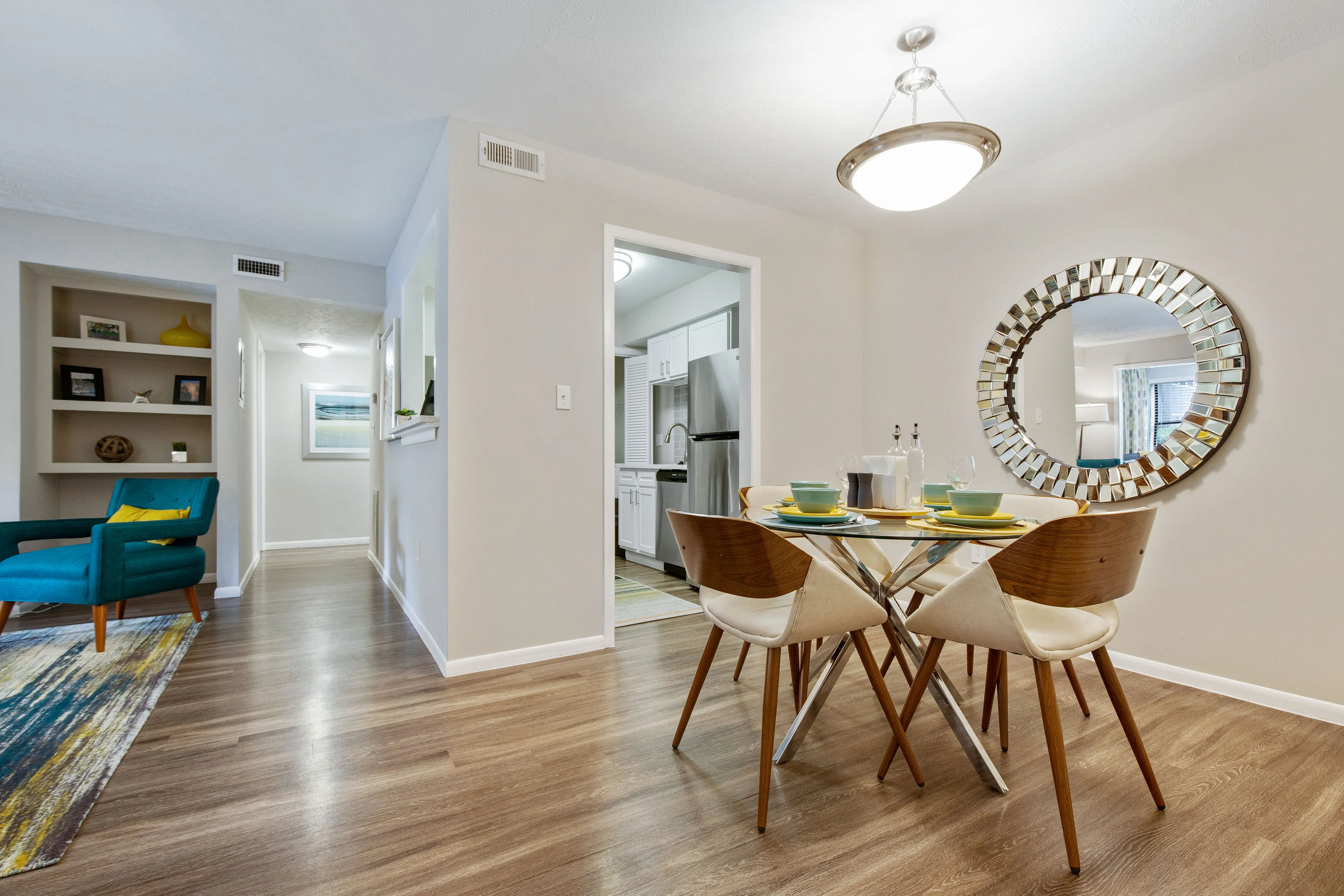 Modern open-concept dining area with elegant glass table, stylish chairs, and a large decorative mirror, leading to a sleek kitchen.