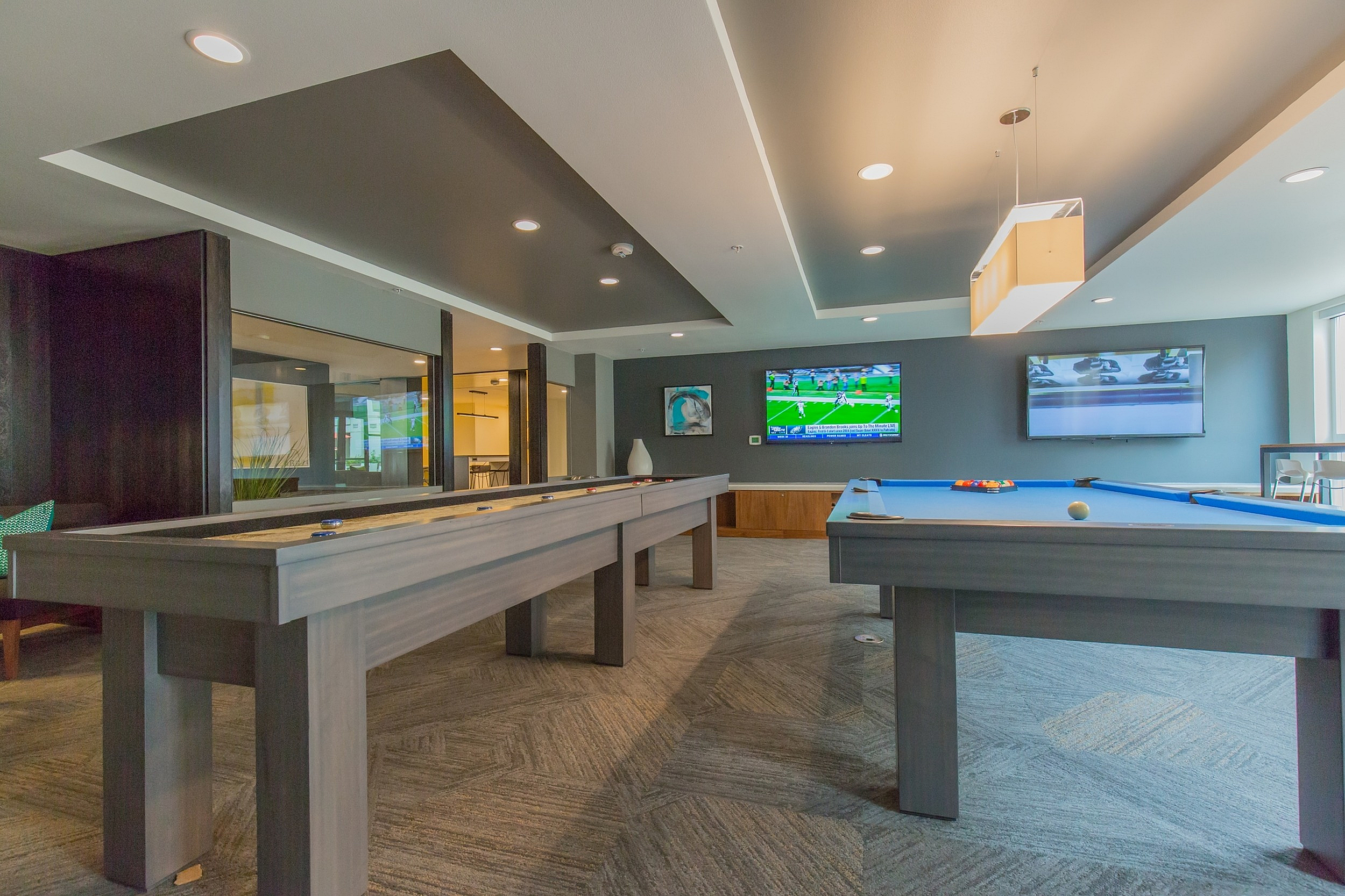 Modern game room with elegant pool and shuffleboard tables, ambient lighting, and multiple TVs for sports entertainment.