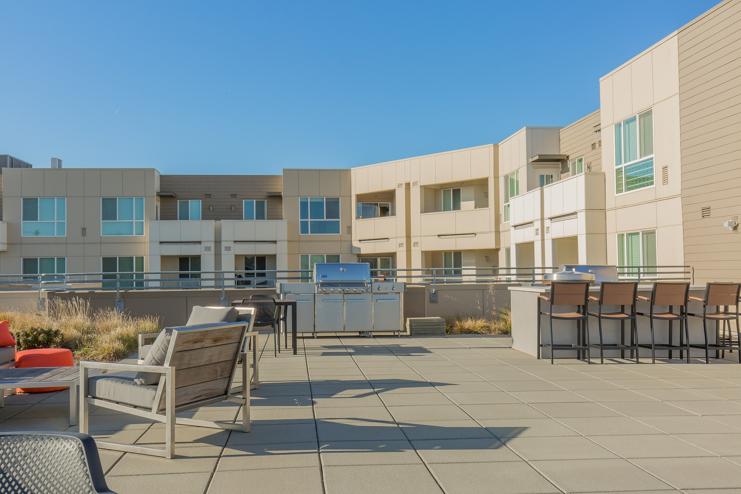 Modern outdoor patio space with stylish seating, BBQ area, and stunning apartment backdrop under a clear blue sky.