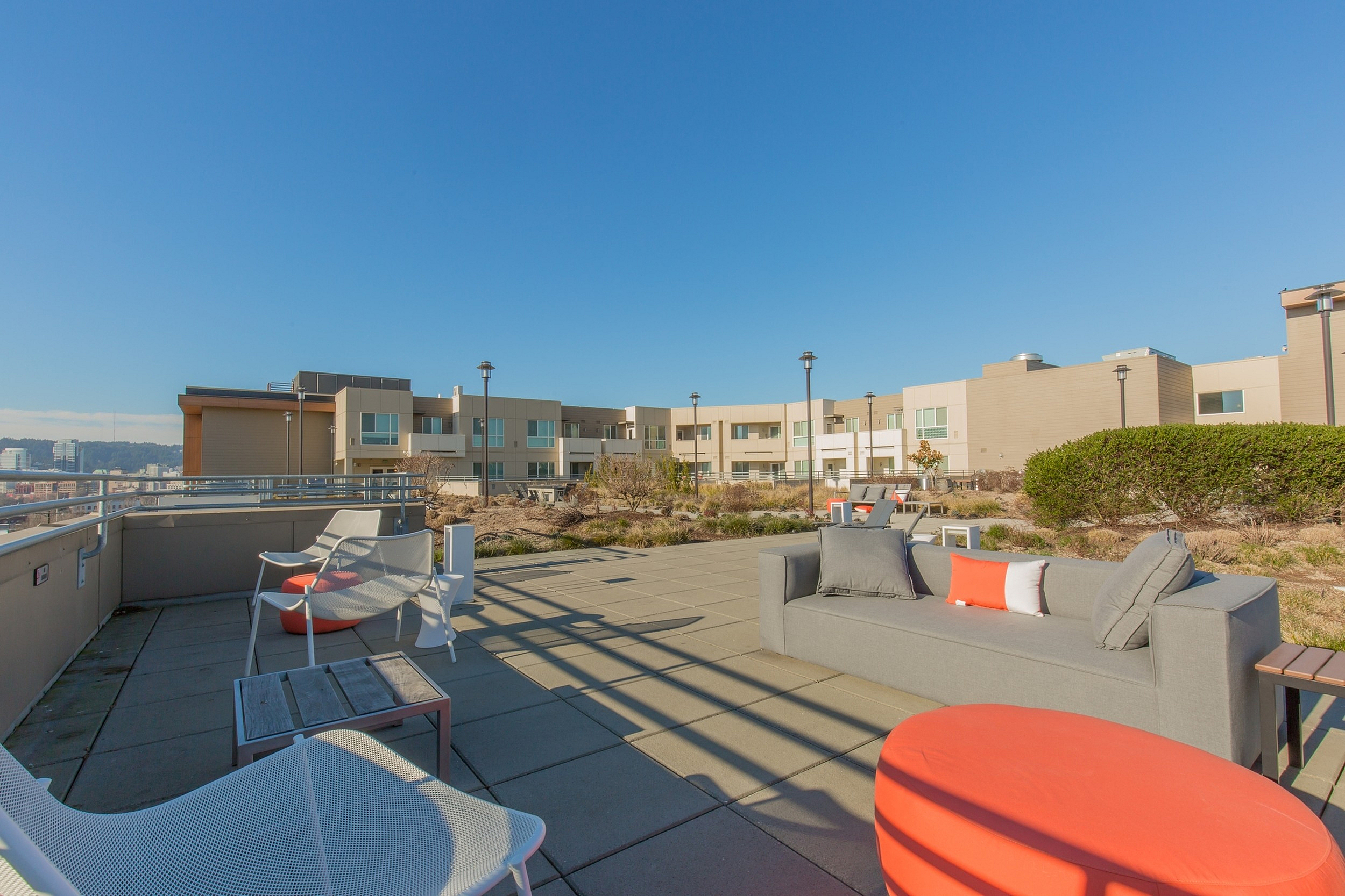 Modern apartment rooftop terrace with stylish seating, vibrant orange accents, and panoramic city views under a clear blue sky.