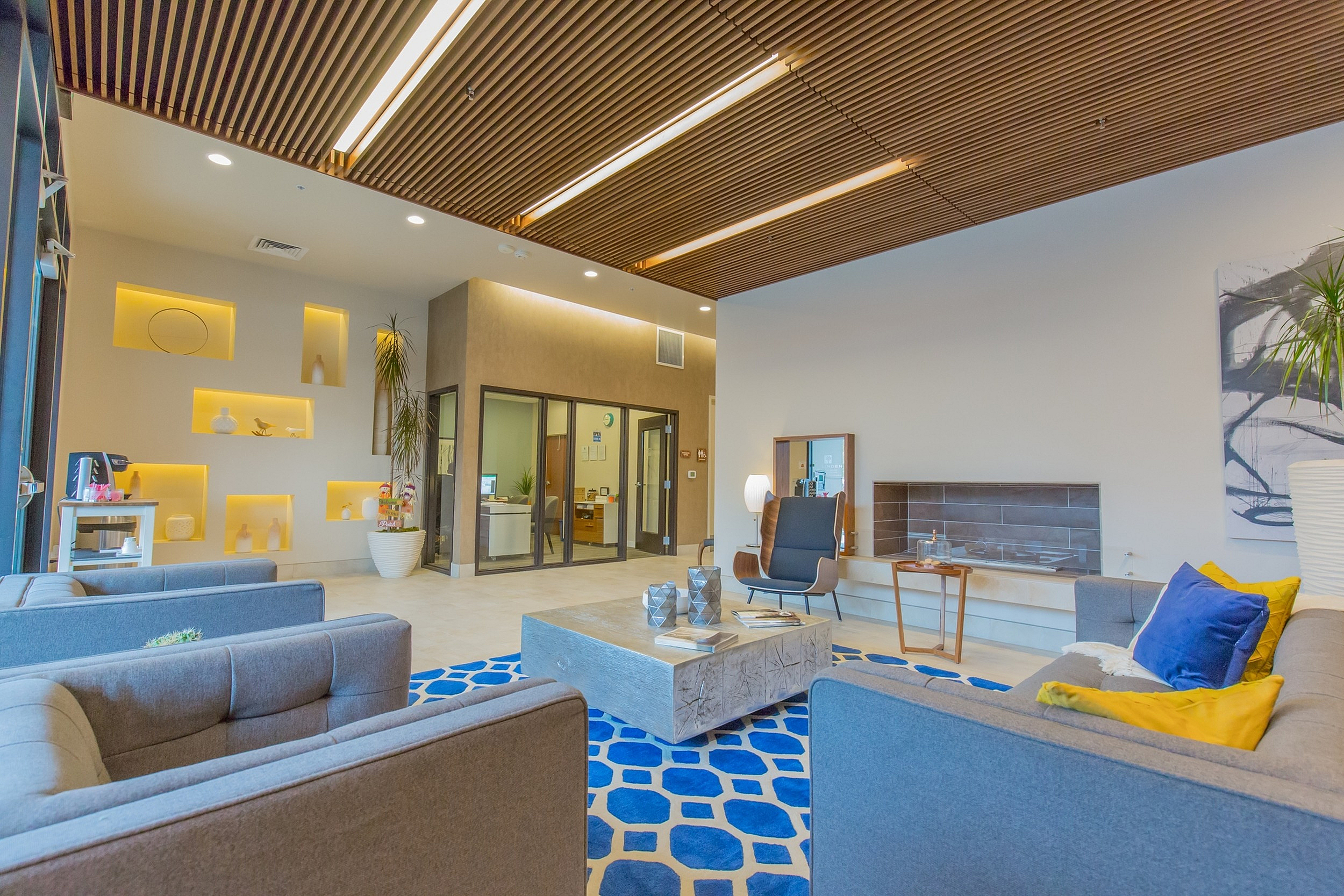 Modern apartment lobby with stylish gray sofas, vibrant blue geometric rug, and sleek wooden ceiling accents.