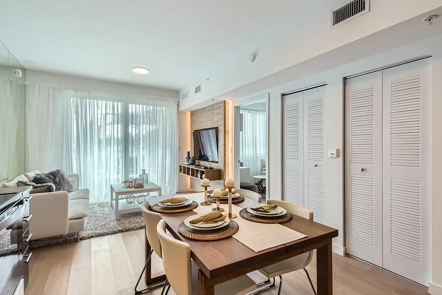 Modern living and dining area with elegant decor, featuring a cozy sofa, dining table set for four, and large windows with sheer curtains.