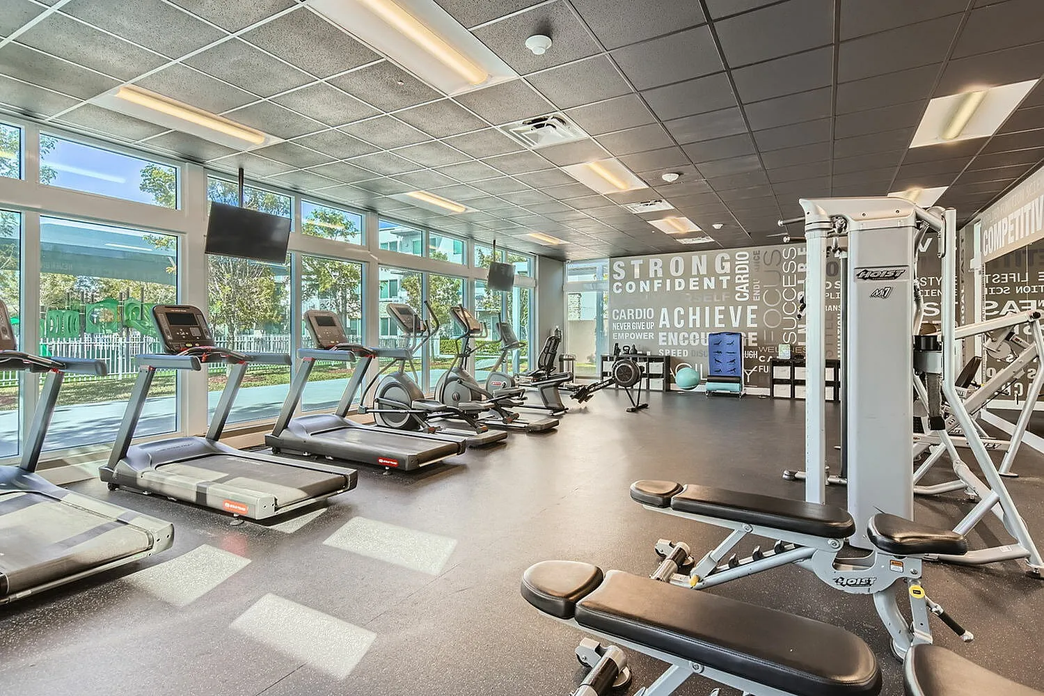 Modern fitness center with large windows, treadmills, exercise equipment, and motivational wall graphics.