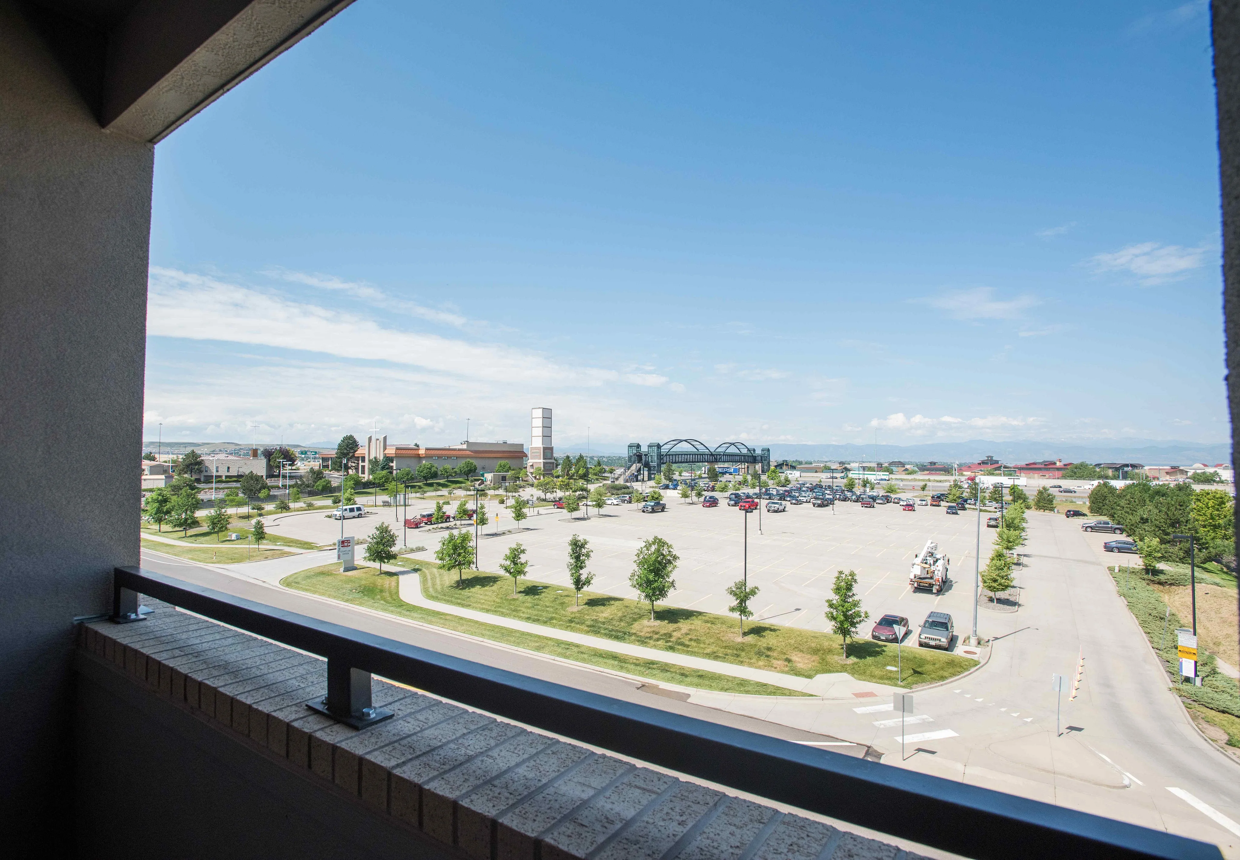 Stunning balcony view featuring a lively commercial area with greenery, parking, and a beautiful horizon under a clear blue sky.