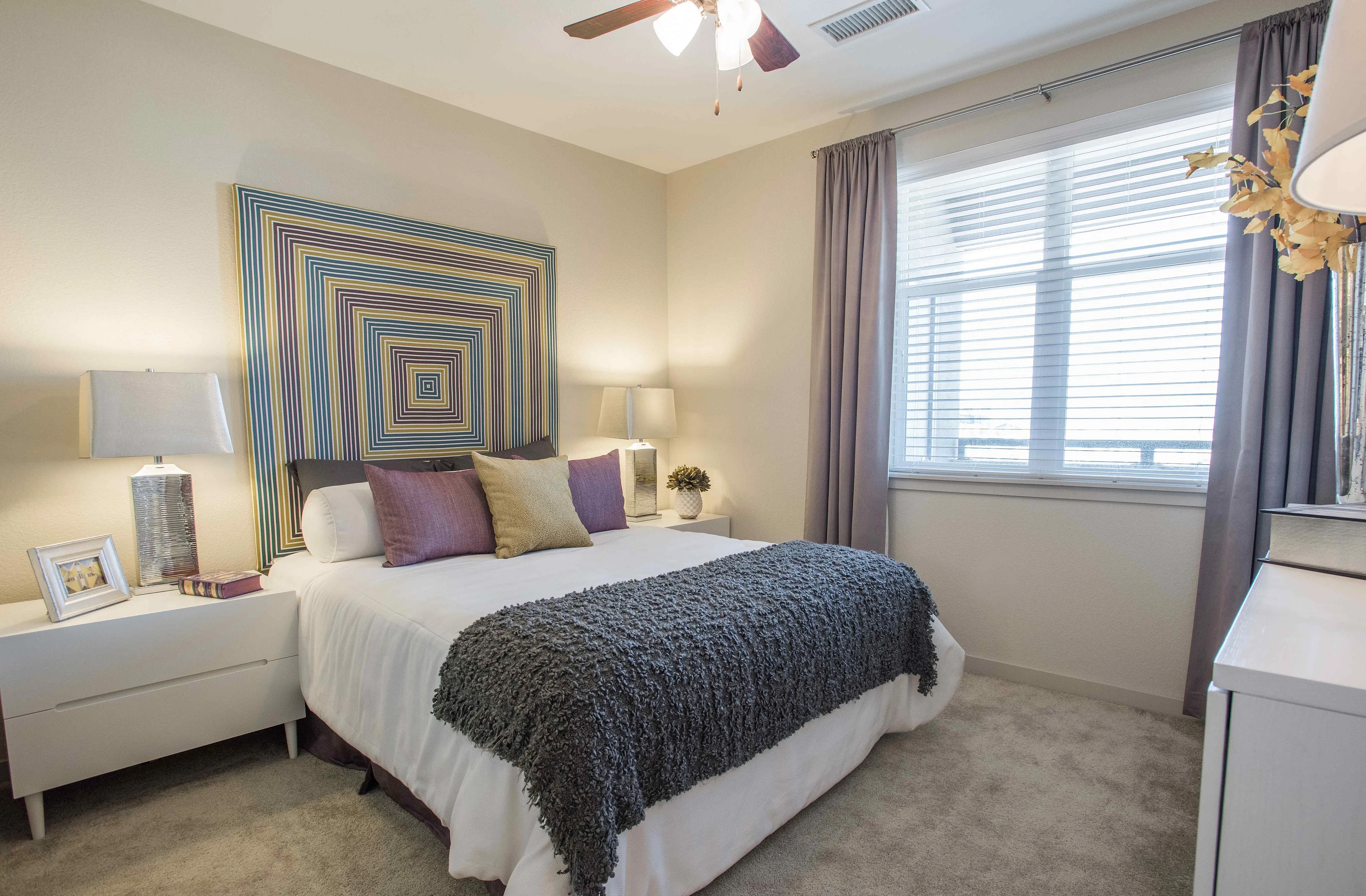 Inviting bedroom with elegant decor, featuring modern art, plush pillows, and ample natural light through large windows.
