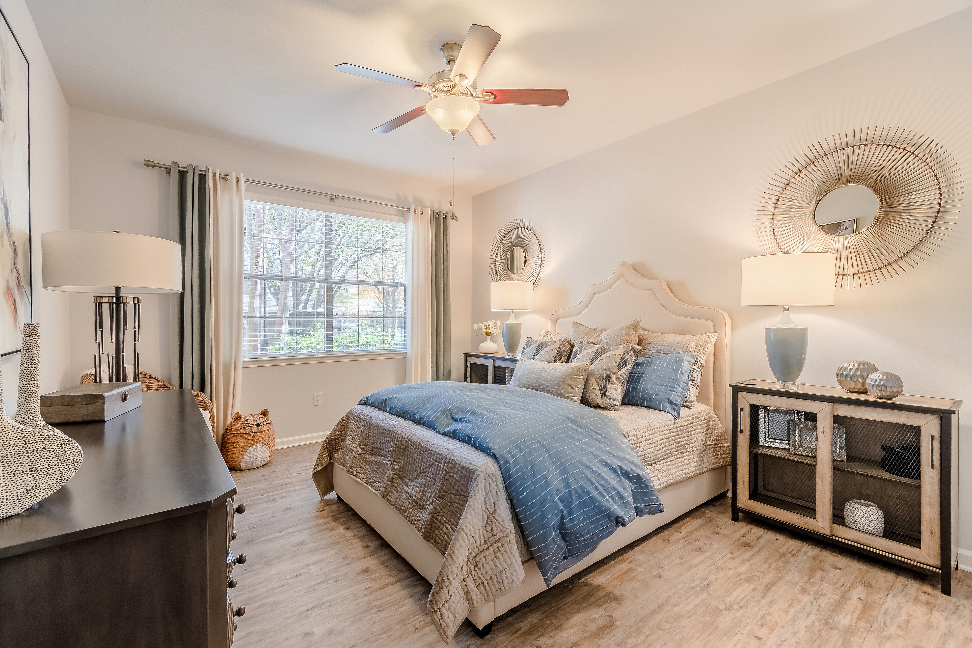 Elegant bedroom featuring a plush bed with blue and neutral linens, decorative mirrors, chic lamps, and a large window inviting natural light, all set on stylish wooden flooring.