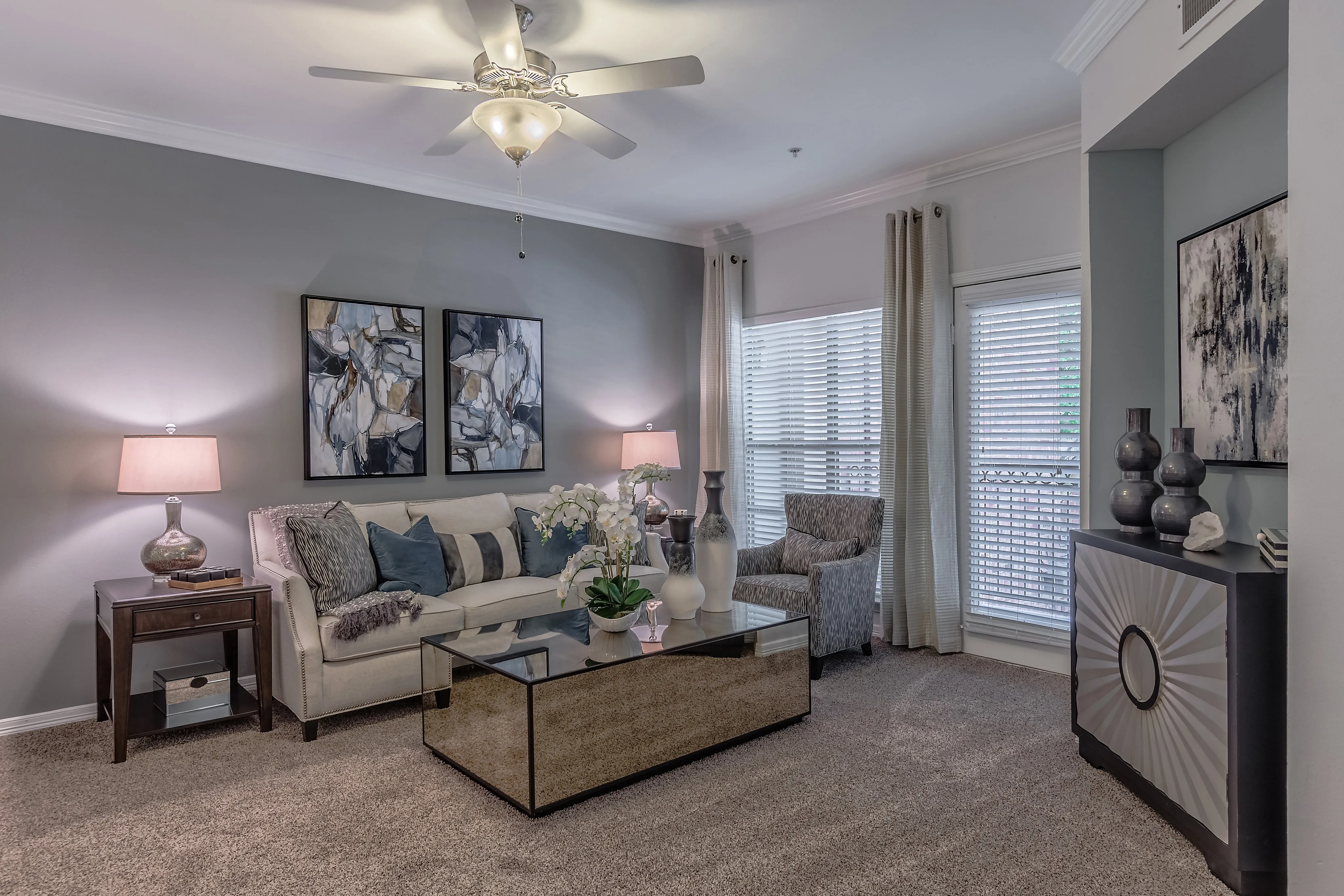 Elegant living room with plush beige sofa, abstract artwork, and stylish glass coffee table under a ceiling fan.