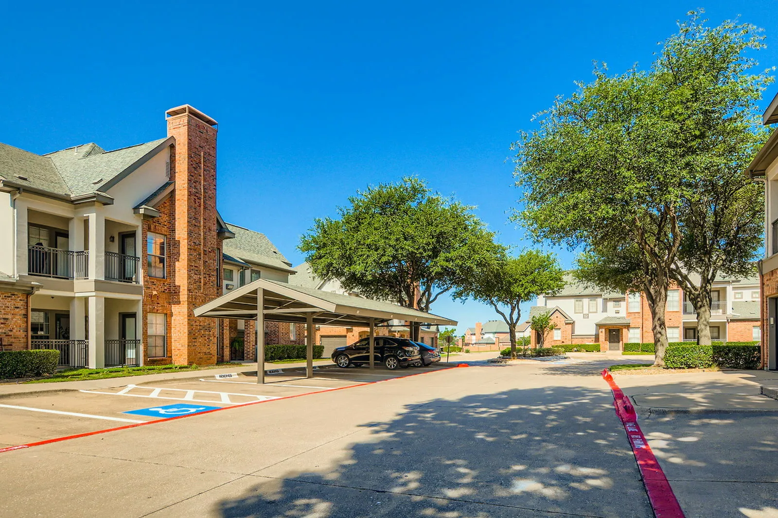Charming apartment complex with elegant brick facades, lush green trees, and ample covered parking under a clear blue sky.