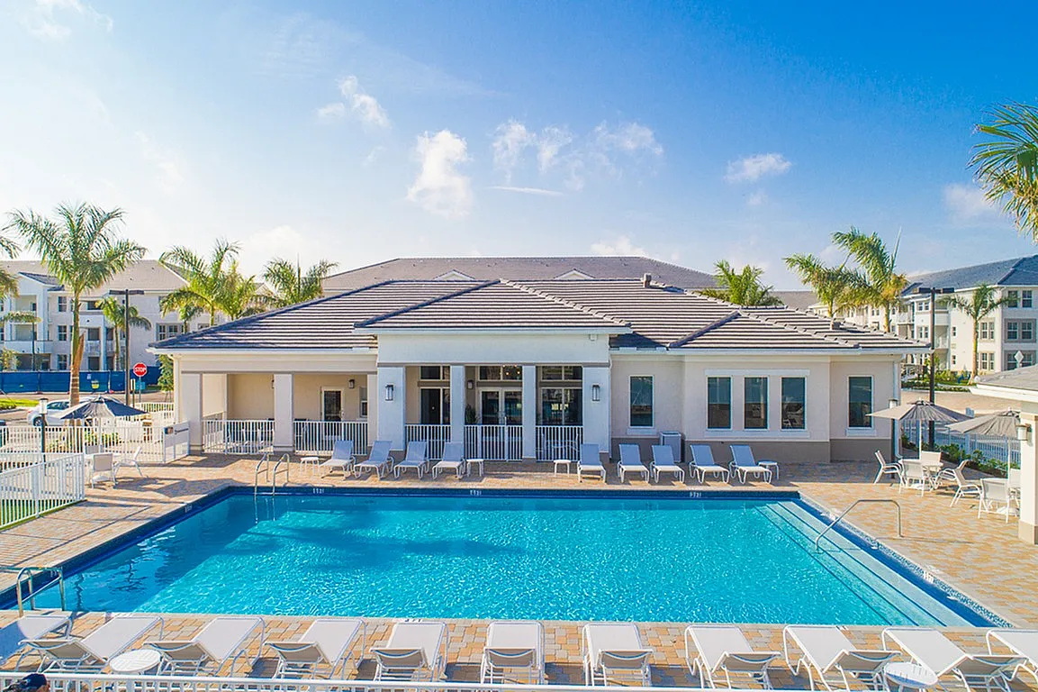 Luxurious apartment complex featuring a crystal-clear swimming pool, surrounded by elegant lounge chairs and tropical palm trees, under a vibrant blue sky.