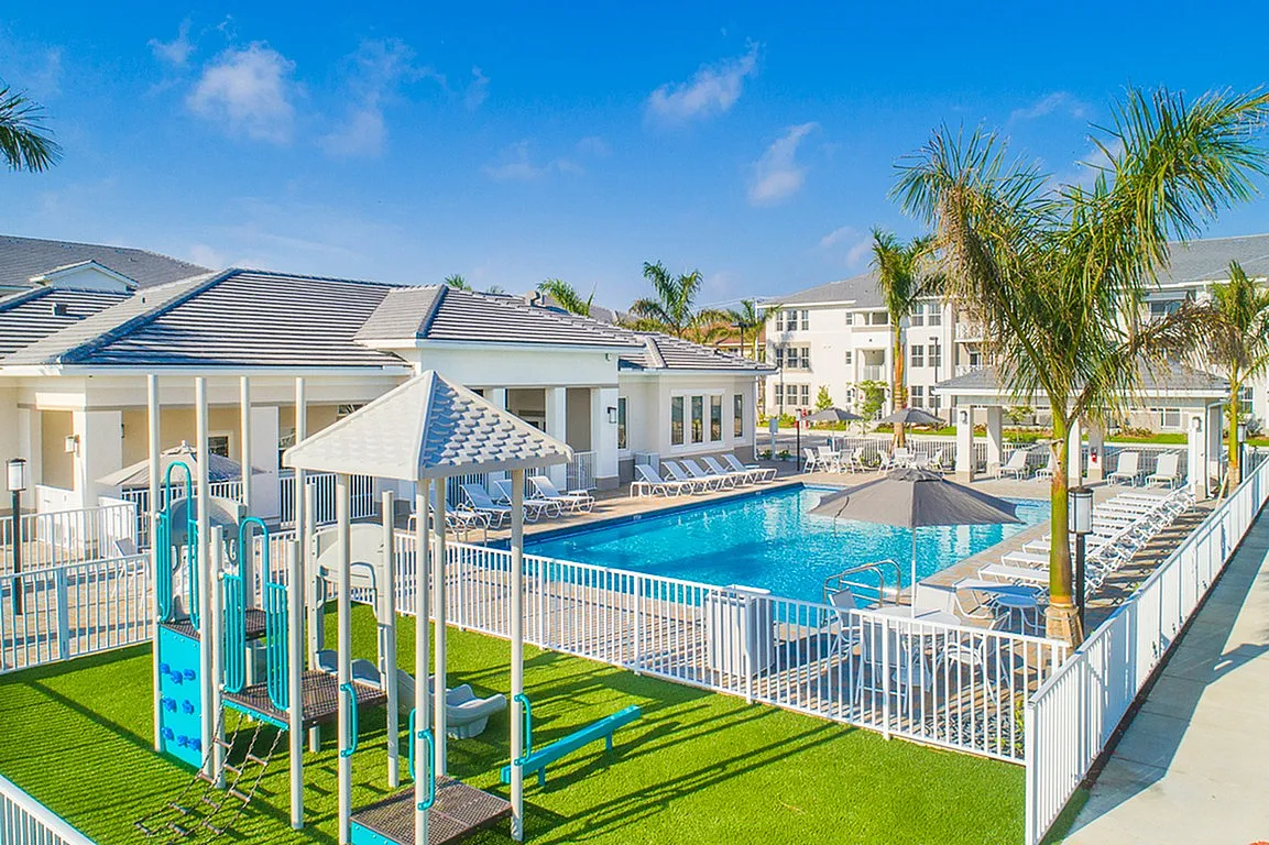 Luxurious apartment complex showcasing a sparkling pool, inviting lounge chairs, and a vibrant playground, surrounded by palm trees under a clear blue sky.
