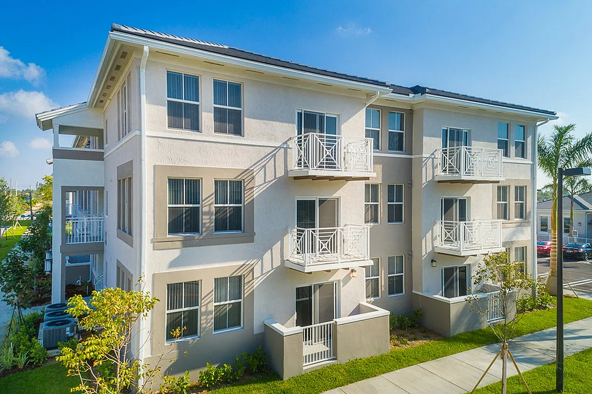 Modern three-story apartment building with pristine white facade, private balconies, and lush landscaping under a clear blue sky.