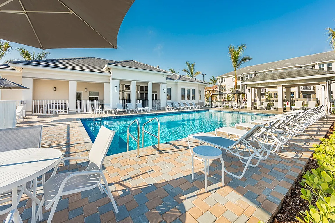 Luxurious poolside area with elegant sun loungers and umbrellas, complete with a stunning modern clubhouse backdrop under a clear blue sky.