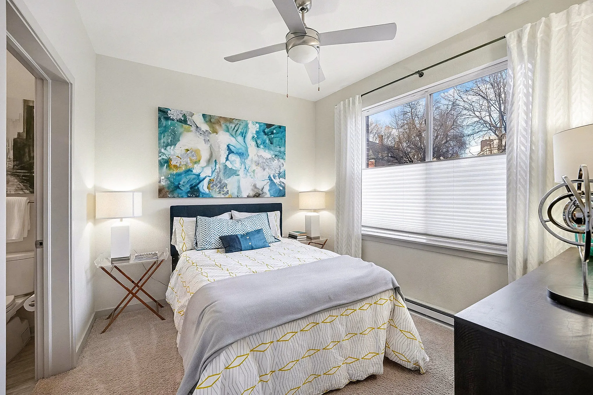 Inviting bedroom with modern decor, featuring a cozy bed, abstract art, and natural light through large windows.