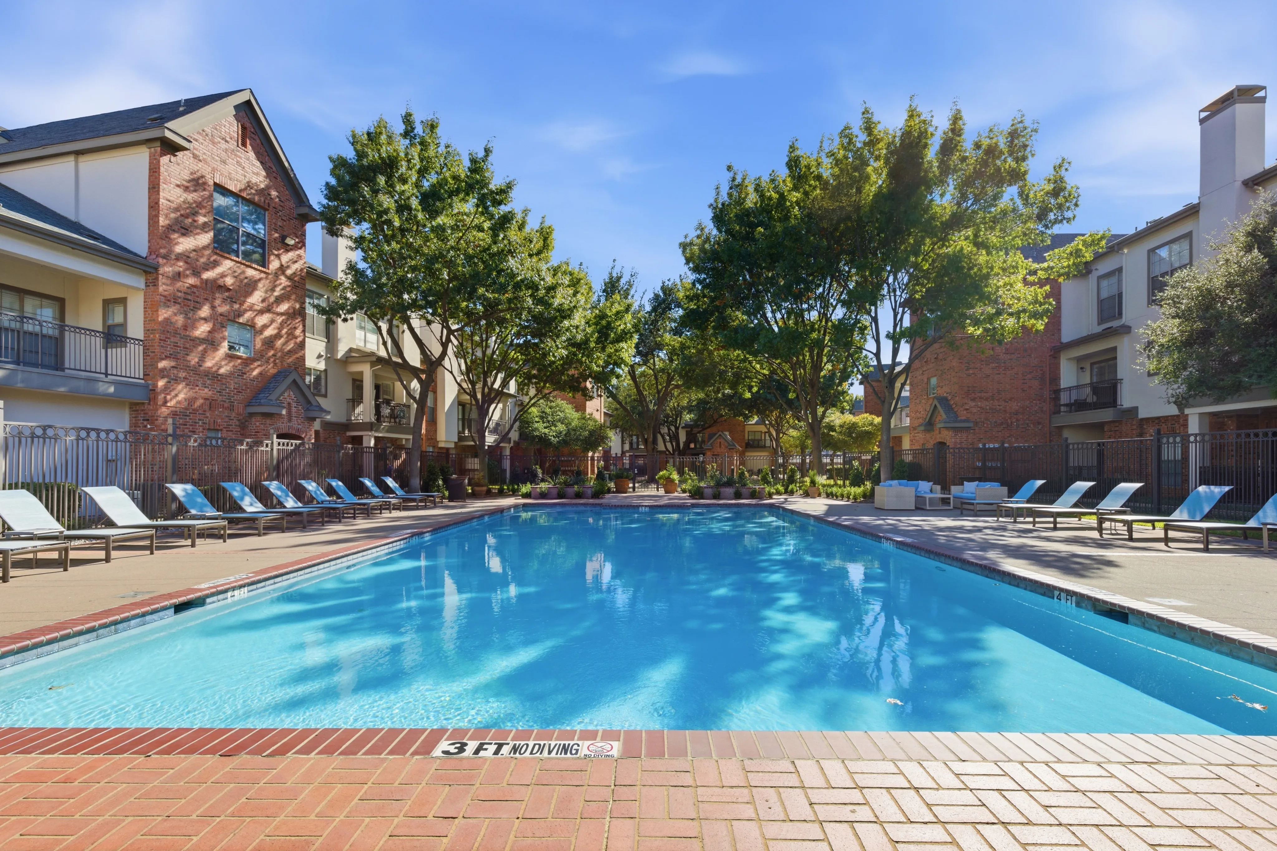Luxurious apartment complex with a sparkling pool, surrounded by lush greenery and elegant brick architecture.