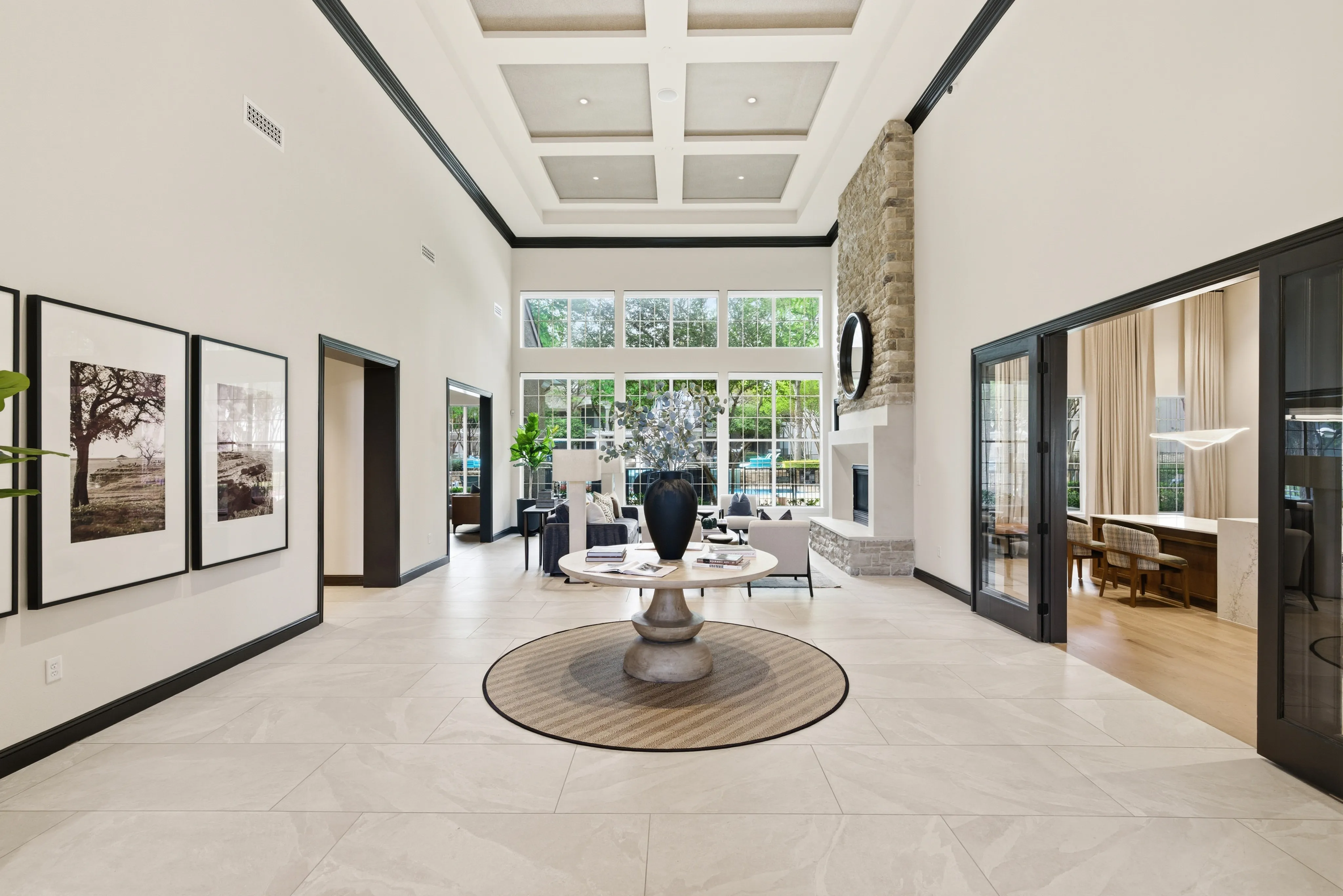Elegant living area with high ceilings, grand chandelier, and stone fireplace. Large windows illuminate the stylish decor, creating a welcoming ambiance.