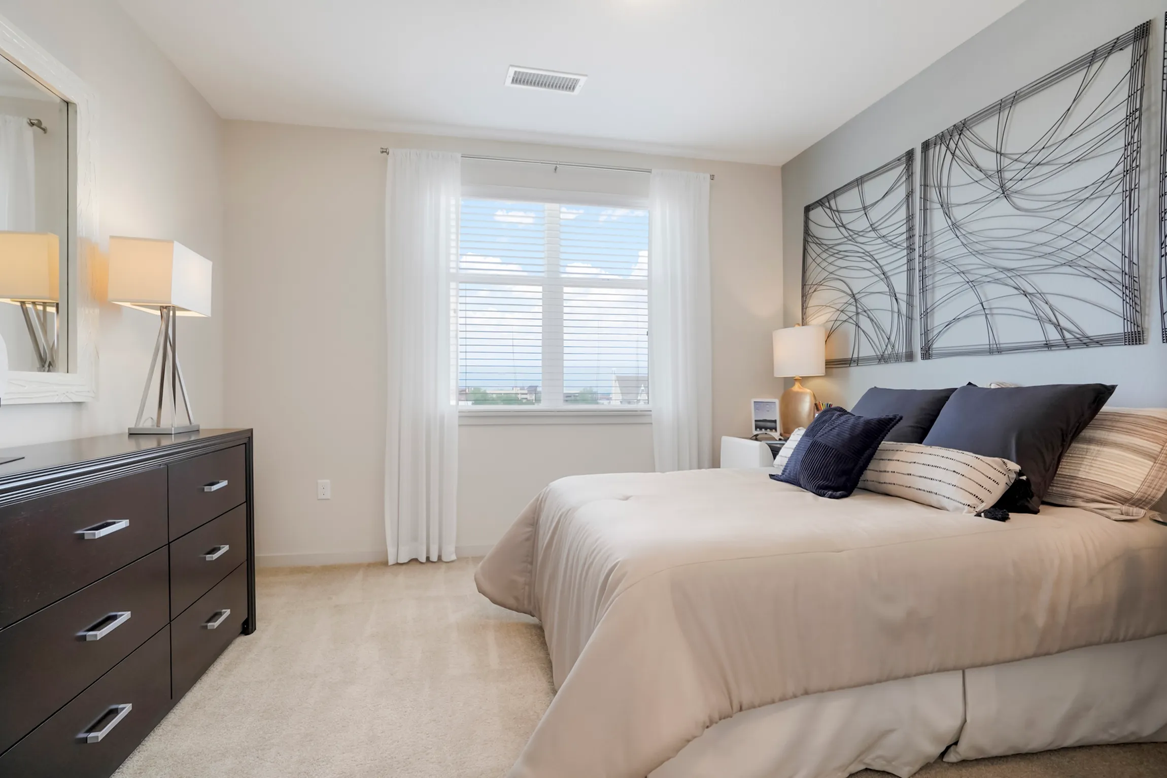 Inviting bedroom with elegant decor, featuring modern art, plush pillows, and ample natural light through large windows.