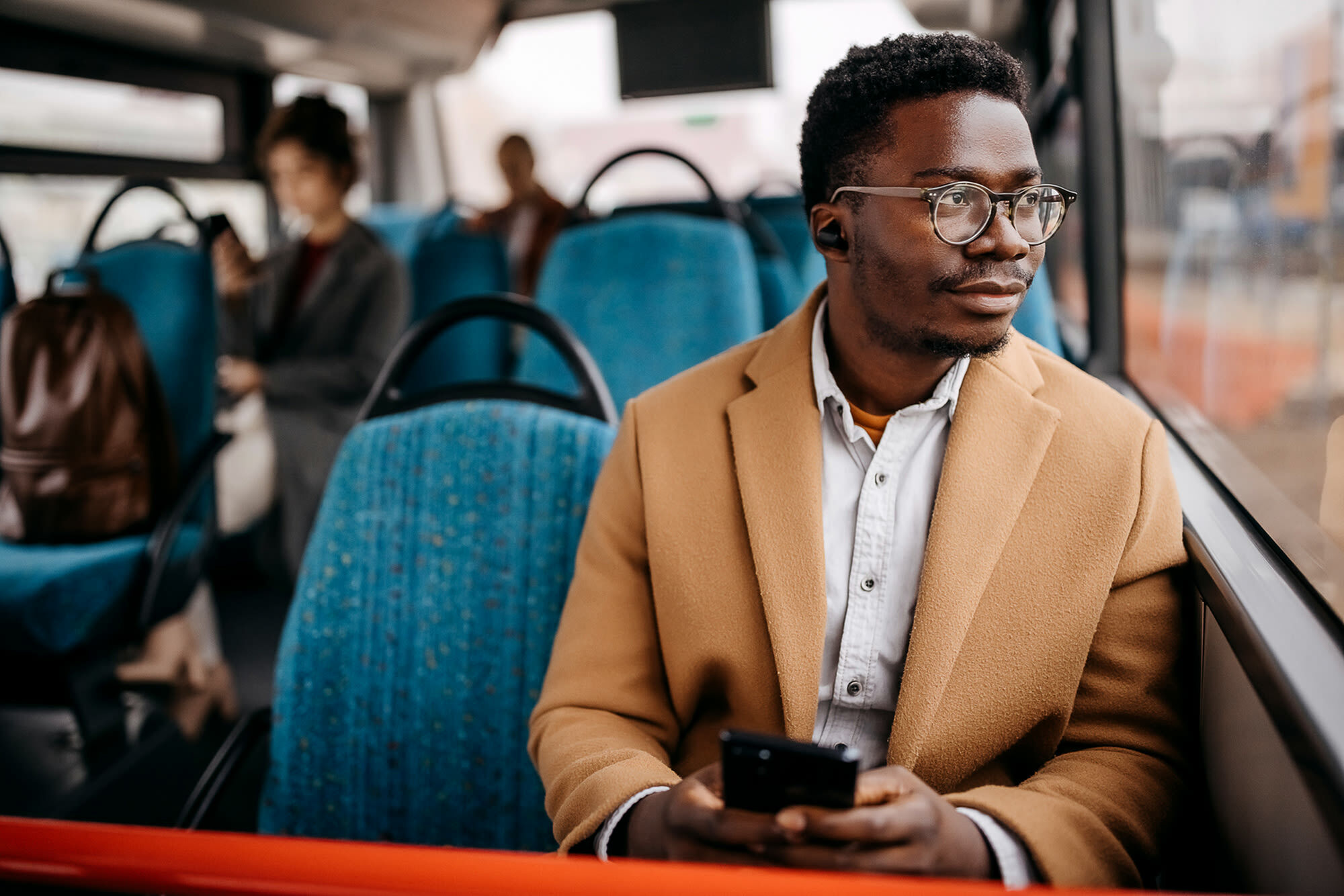 Man wearing glasses and a beige coat, enjoying a peaceful bus ride while holding a smartphone, seated by the window.