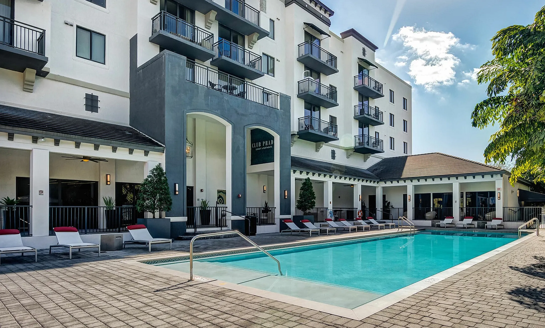 Modern apartment complex with elegant architecture, featuring a sparkling swimming pool and relaxing lounge area under clear skies.