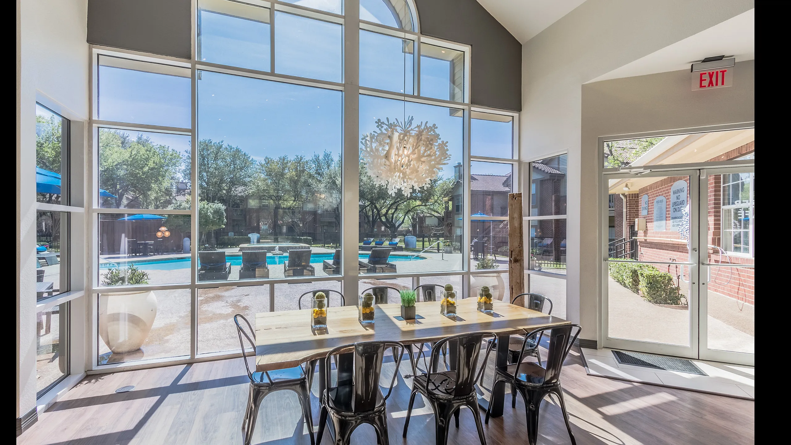 Bright and inviting dining area with a large table, trendy metal chairs, and a stunning poolside view through expansive windows.