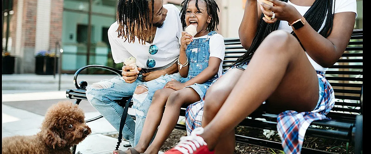 Joyful family moment as a father, mother, and daughter enjoy ice cream together on a sunny day, accompanied by their cute poodle, outside a modern building.