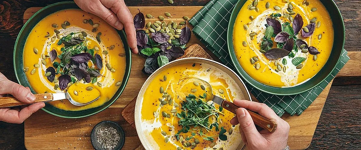 Vibrant autumnal soup bowls garnished with fresh herbs, creamy swirls, and pumpkin seeds, captured over a rustic wooden table.