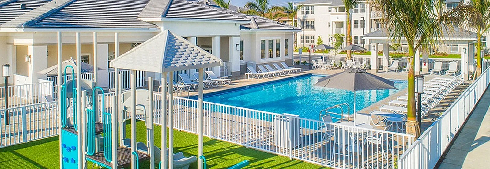 Luxurious apartment complex with a sparkling pool, modern lounge chairs, and a vibrant playground under sunny skies.