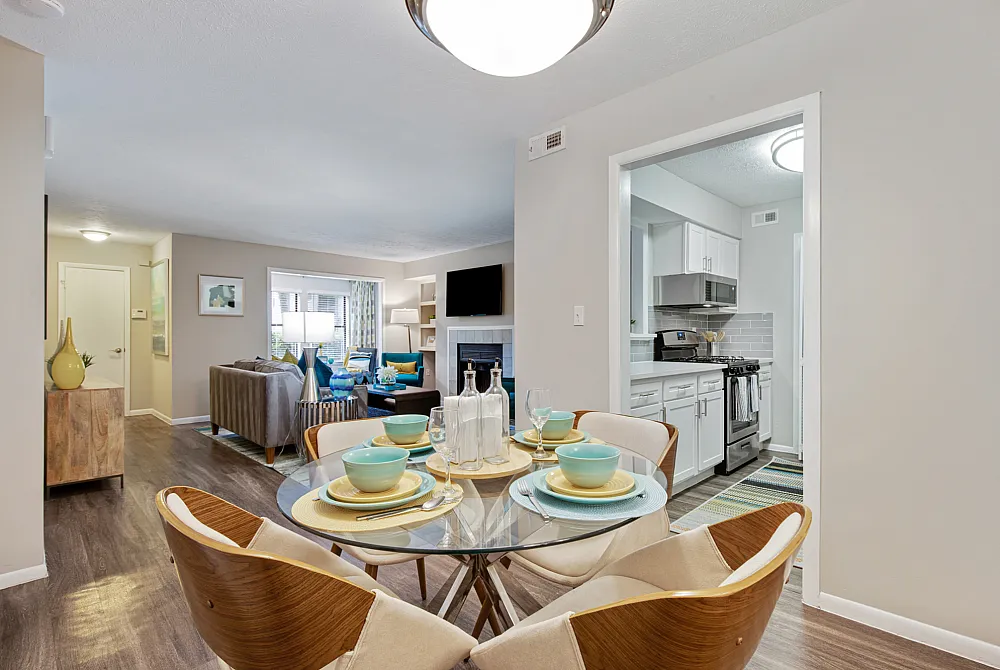 An interior view of a dining area with a table and chairs, a living room area with a couch, flatscreen television, and large window, and partial views of a kitchen at The Madison apartments in Charlotte, North Carolina.