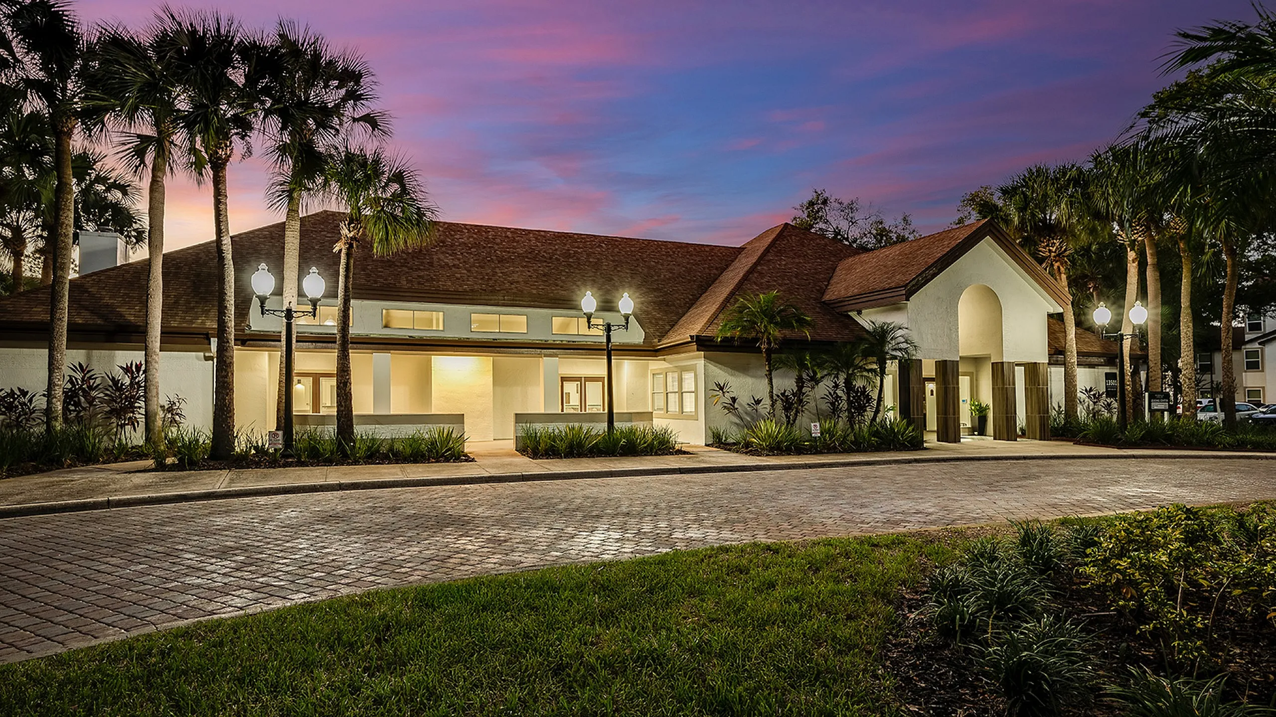 Elegant entrance to a luxurious apartment complex, featuring palm trees and a charming cobblestone driveway under a clear blue sky.