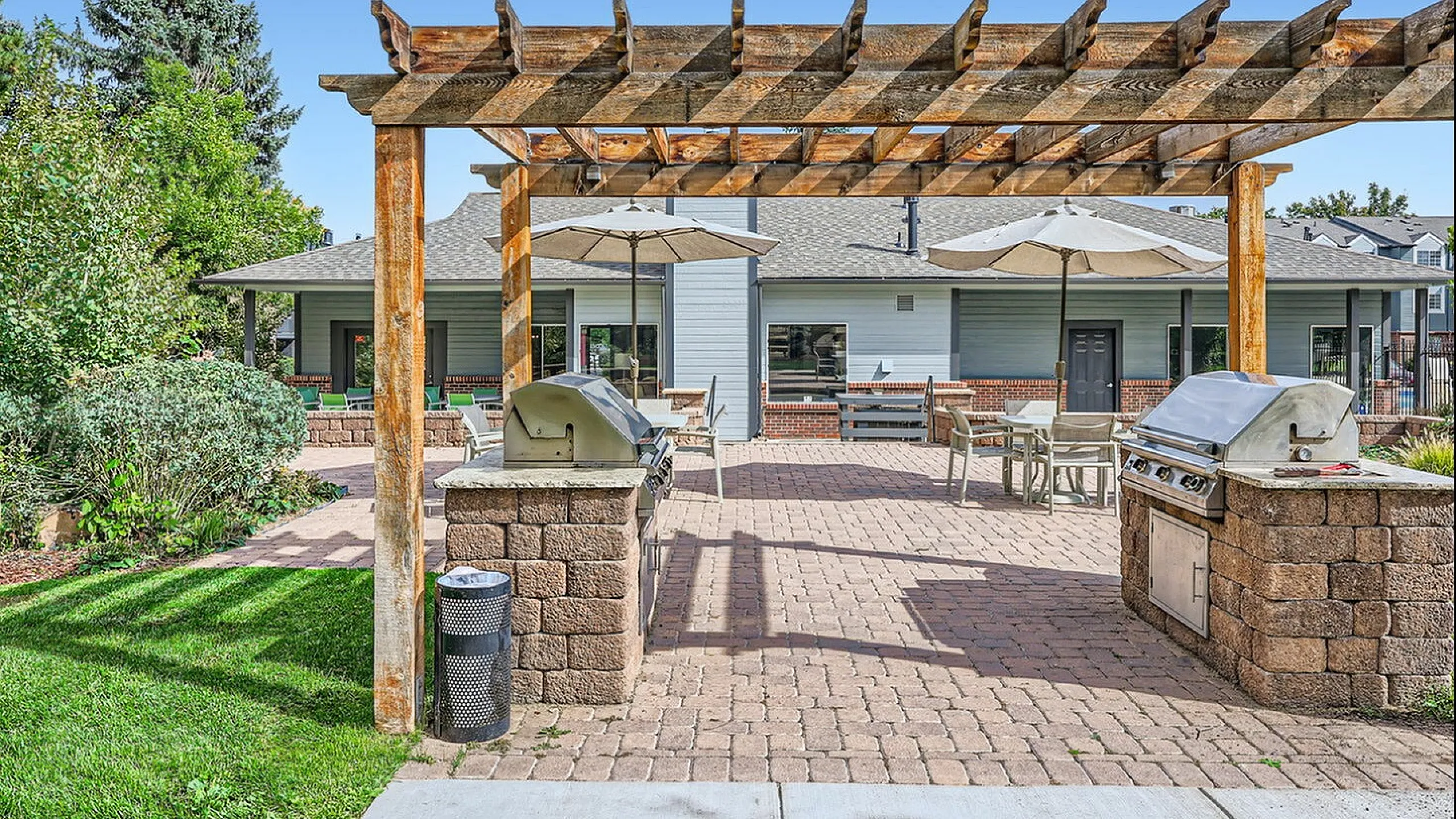 Welcoming outdoor patio area with stylish dining setup under large umbrellas, surrounded by lush greenery and modern apartments in the background.