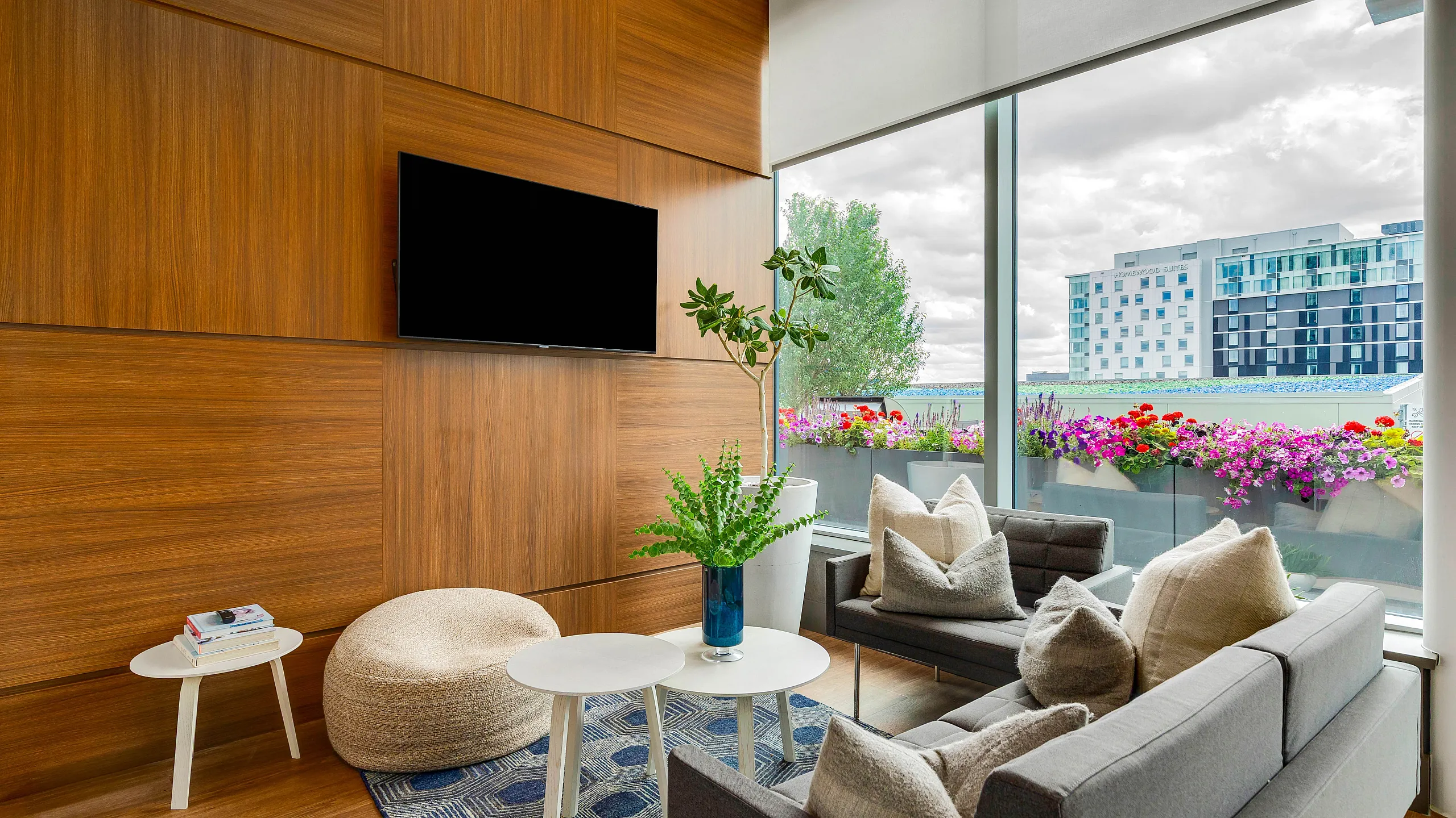 Modern living room with sleek wooden paneling, large windows showcasing a vibrant city view, stylish furniture, and lush greenery.