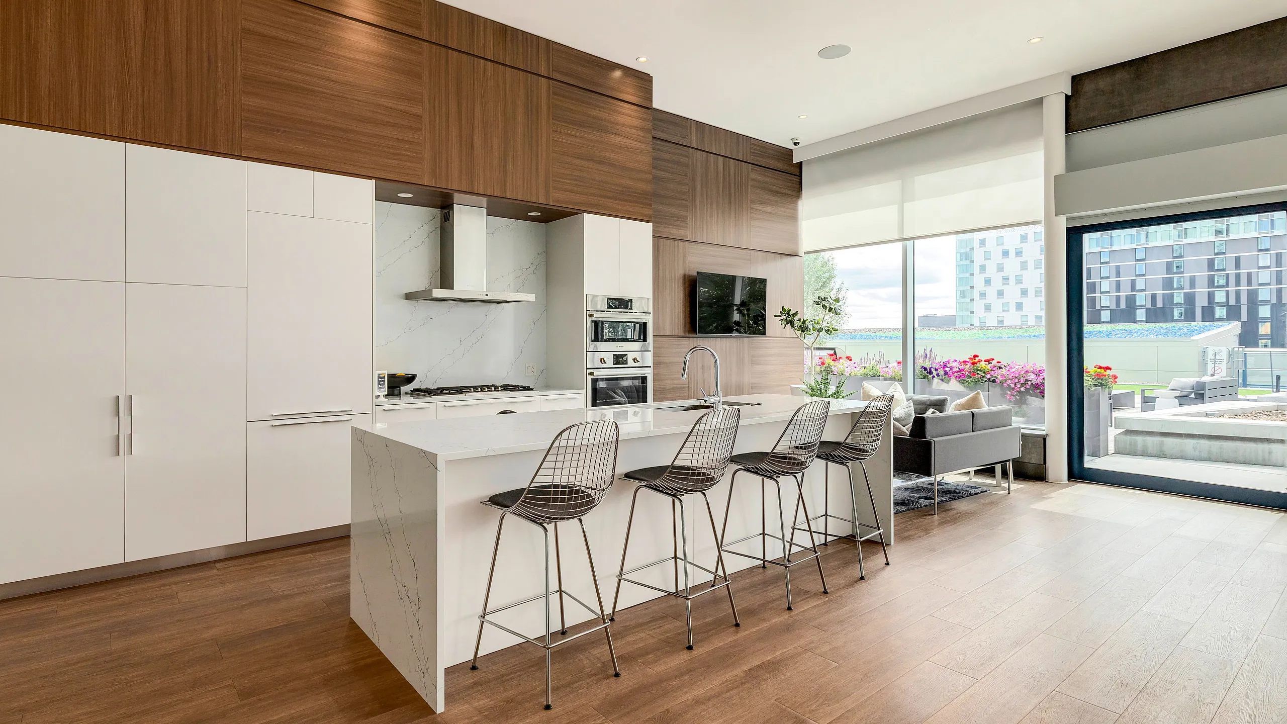 Modern kitchen with sleek white cabinetry, marble countertops, and stainless steel appliances, featuring an inviting breakfast bar with chic wire bar stools, and a view of a sunlit living area with vibrant outdoor greenery.