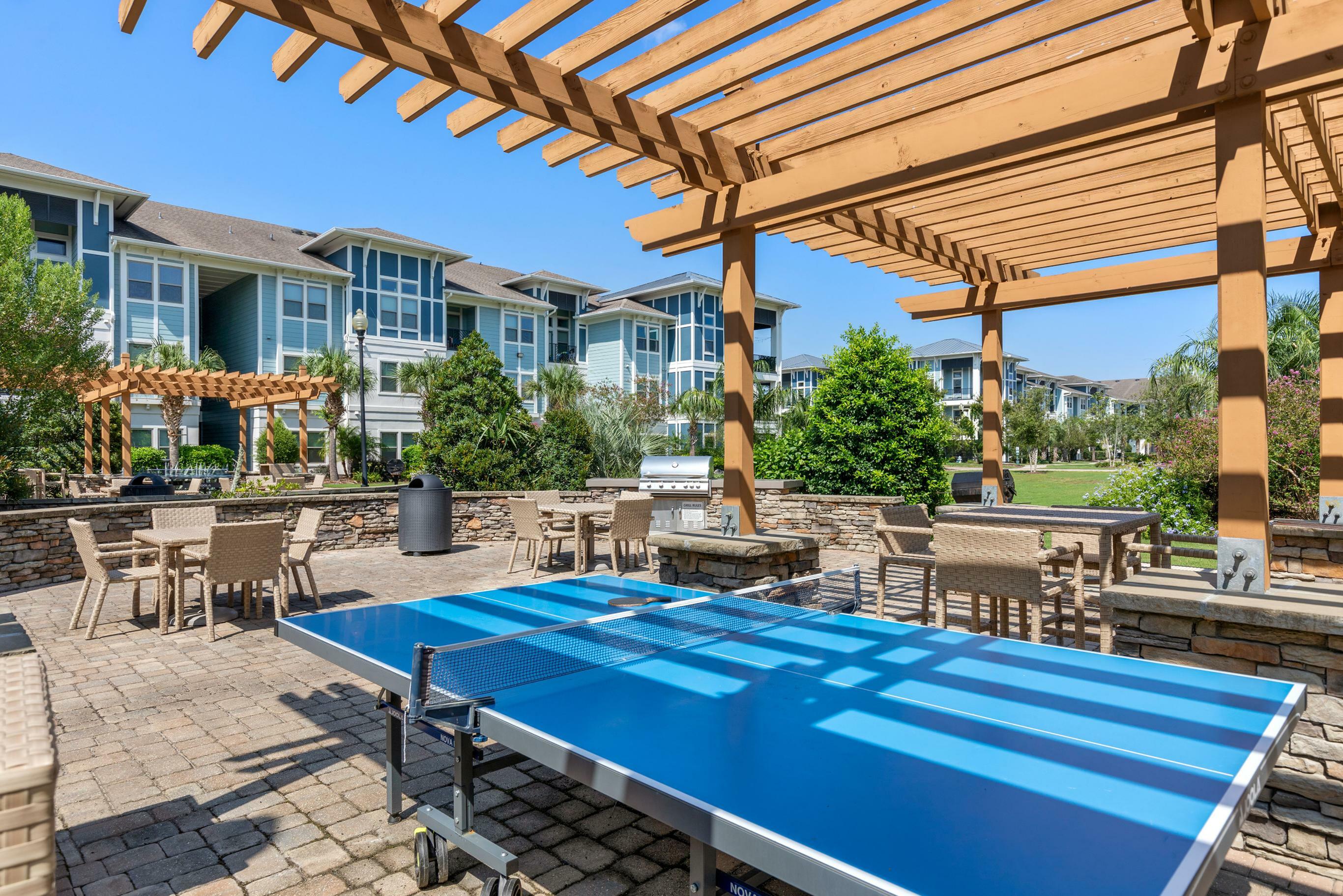 Chic outdoor recreation area featuring a vibrant blue ping pong table under a stylish wooden pergola, with modern patio seating and lush landscaping in the background.