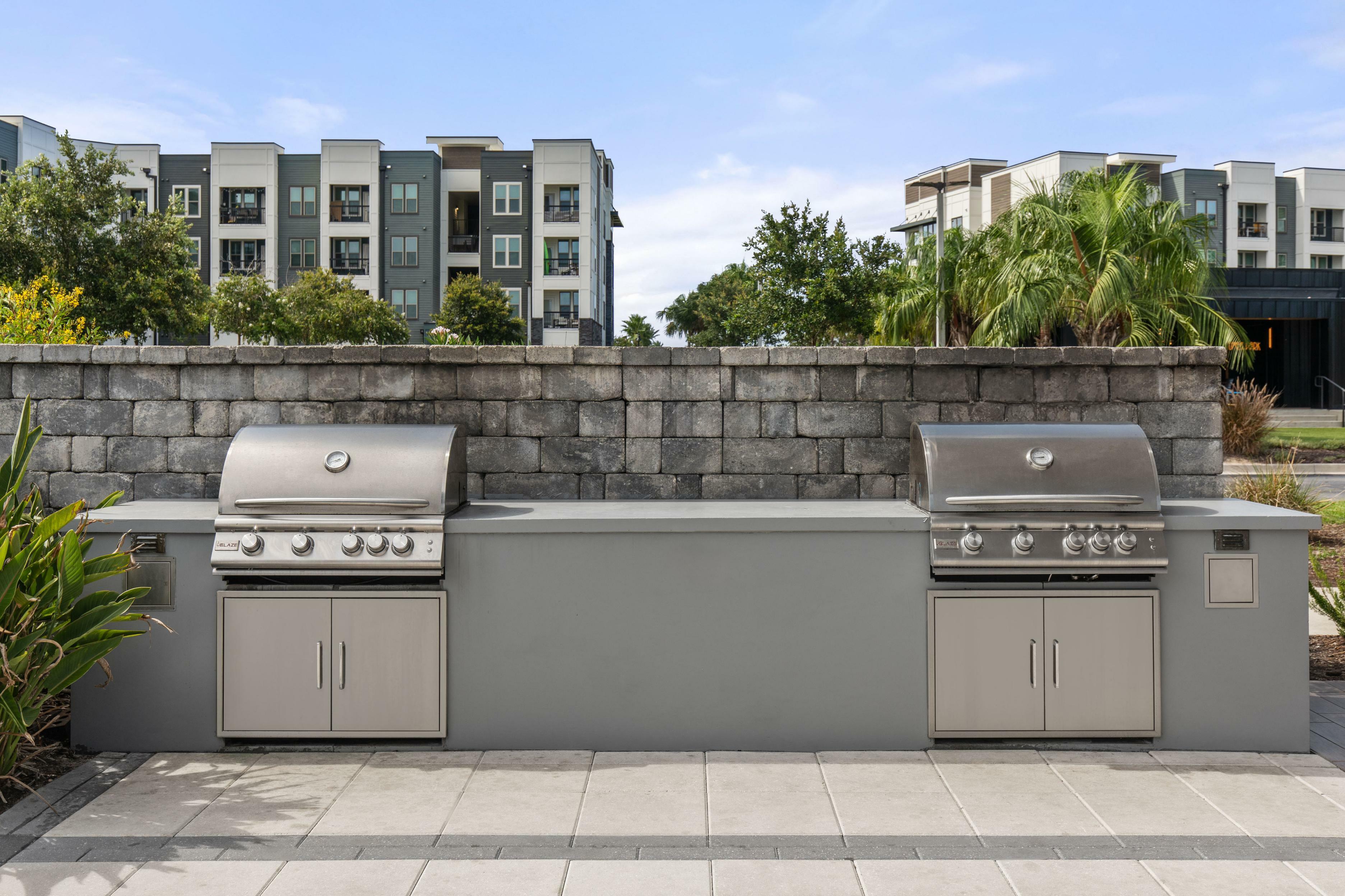 Elegant outdoor grilling station with dual stainless steel barbeques, set against a stylish stone wall backdrop and lush apartment surroundings.