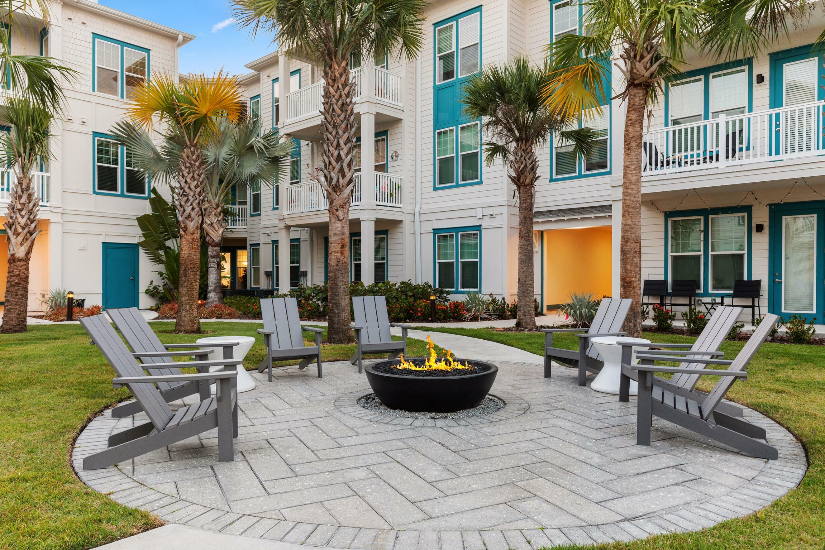 Charming apartment complex courtyard featuring a cozy fire pit surrounded by Adirondack chairs and lush palm trees.