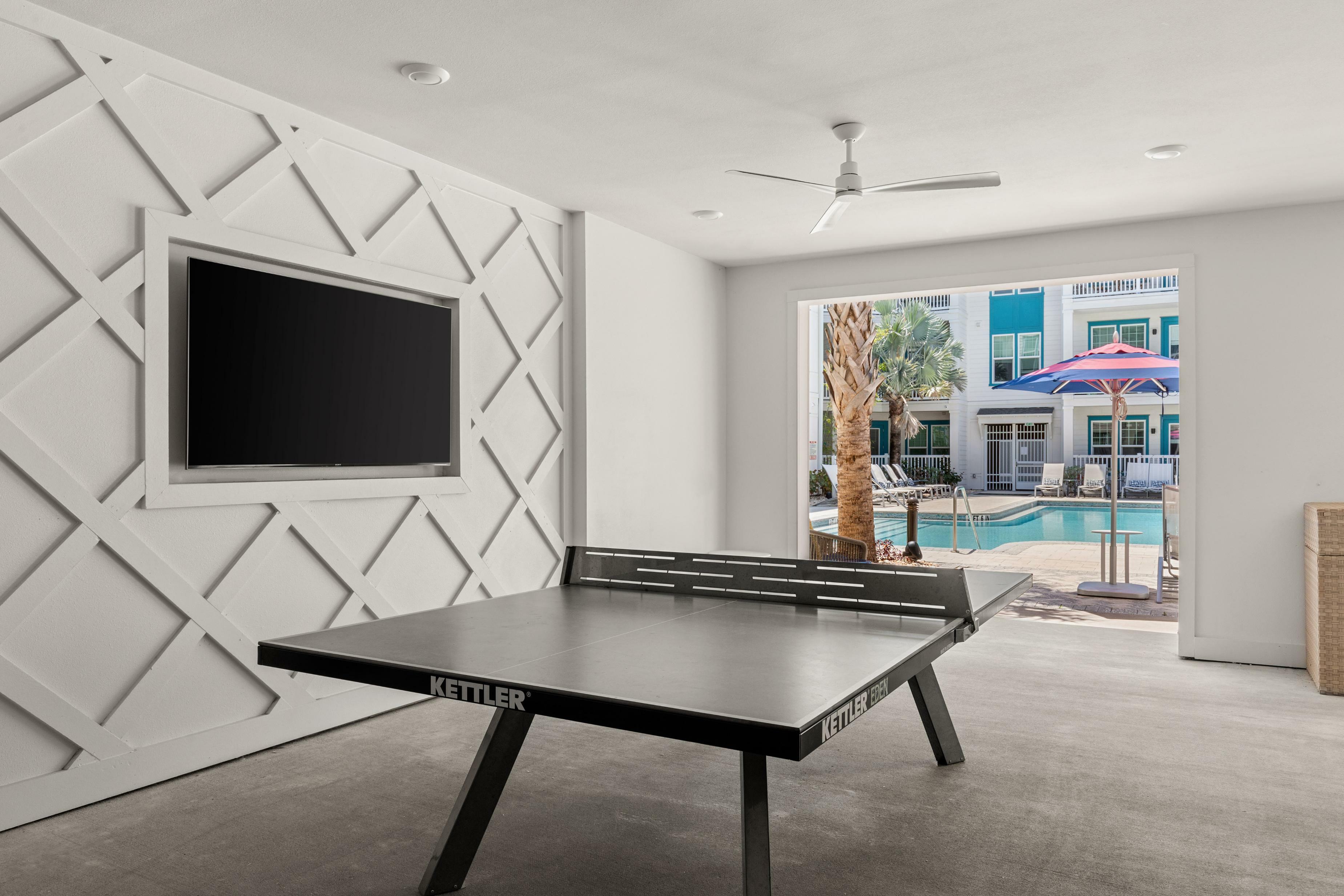 Luxurious recreation room featuring a stylish ping pong table, modern wall design, and a stunning view of the sunlit pool area with palm trees and umbrellas.